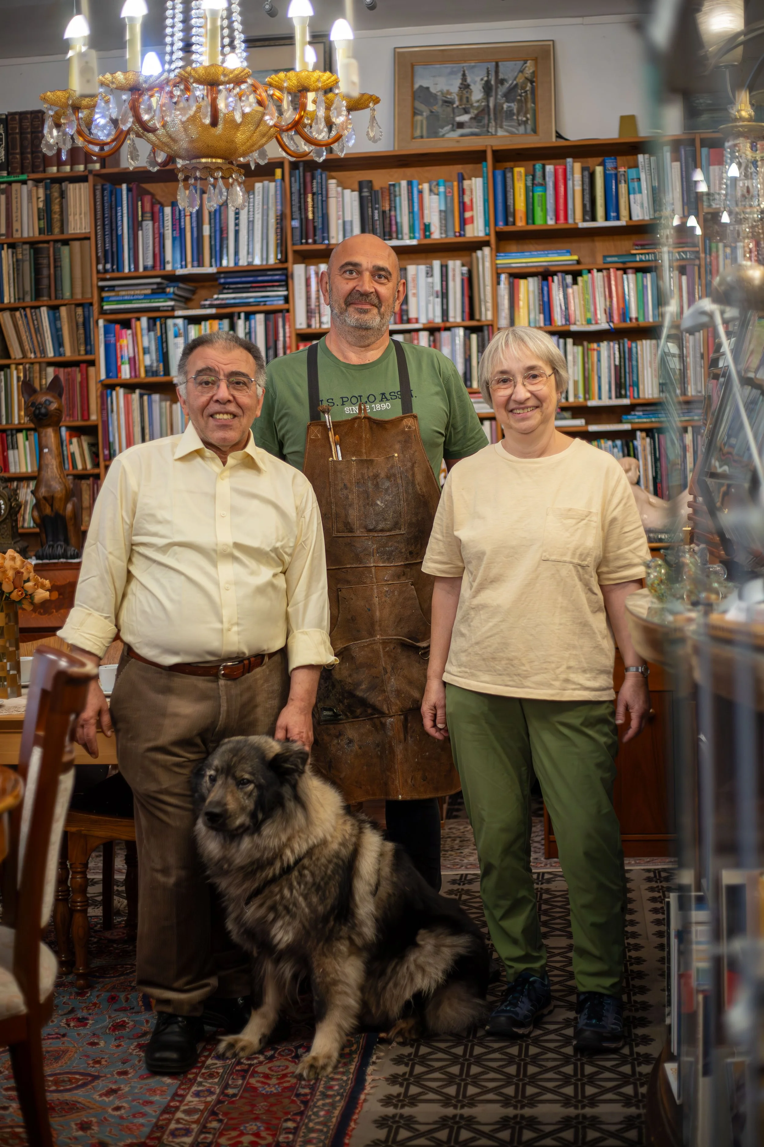 Drei Personen und ein Hund in einer Bücherhandlung. Zwei Frauen, ein Mann mit Bart und Brille und ein großer Hund sitzen und stehen vor einem Bücherregal, im Hintergrund eine Kristalllampe und eine eingerahmte Malerei.