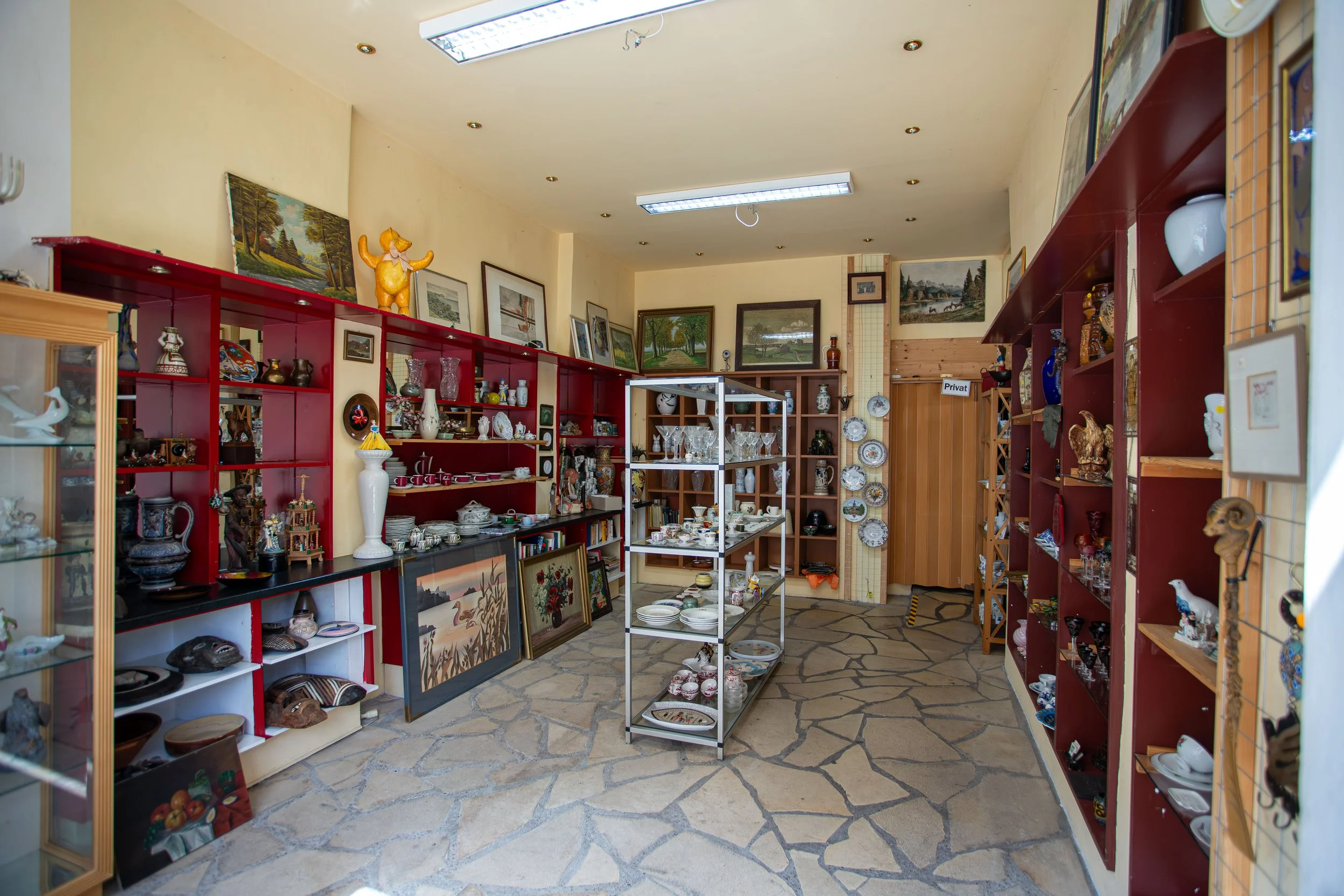 Innenraum eines Secondhand-Ladens mit roten und natürlichen Holzregalen, die verschiedenes Geschirr, Kunstwerke und Dekorationsgegenstände zeigen. Es liegen gemalte Bilder auf den Regalen. Die Decke hat Einbaulampen und eine verlegte Steinboden. Es gibt ein kleines Metallregal in der Mitte mit Porzellan und Gläsern.