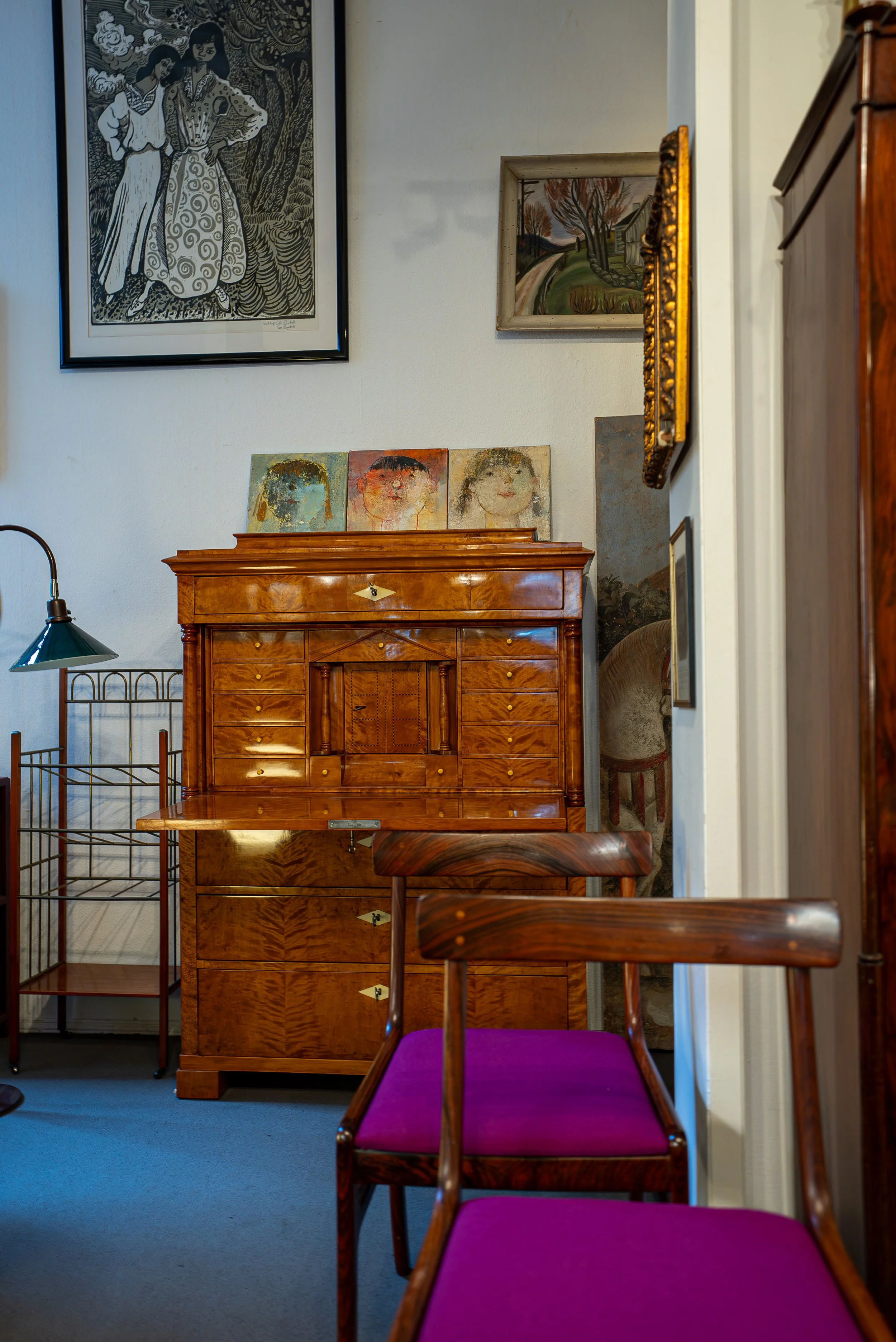 Ein Inneneinrichtung mit einem alten Holzschreibtisch, mehreren bunten Stühlen mit lila Polster, Wandbildern und einem Wandregal.