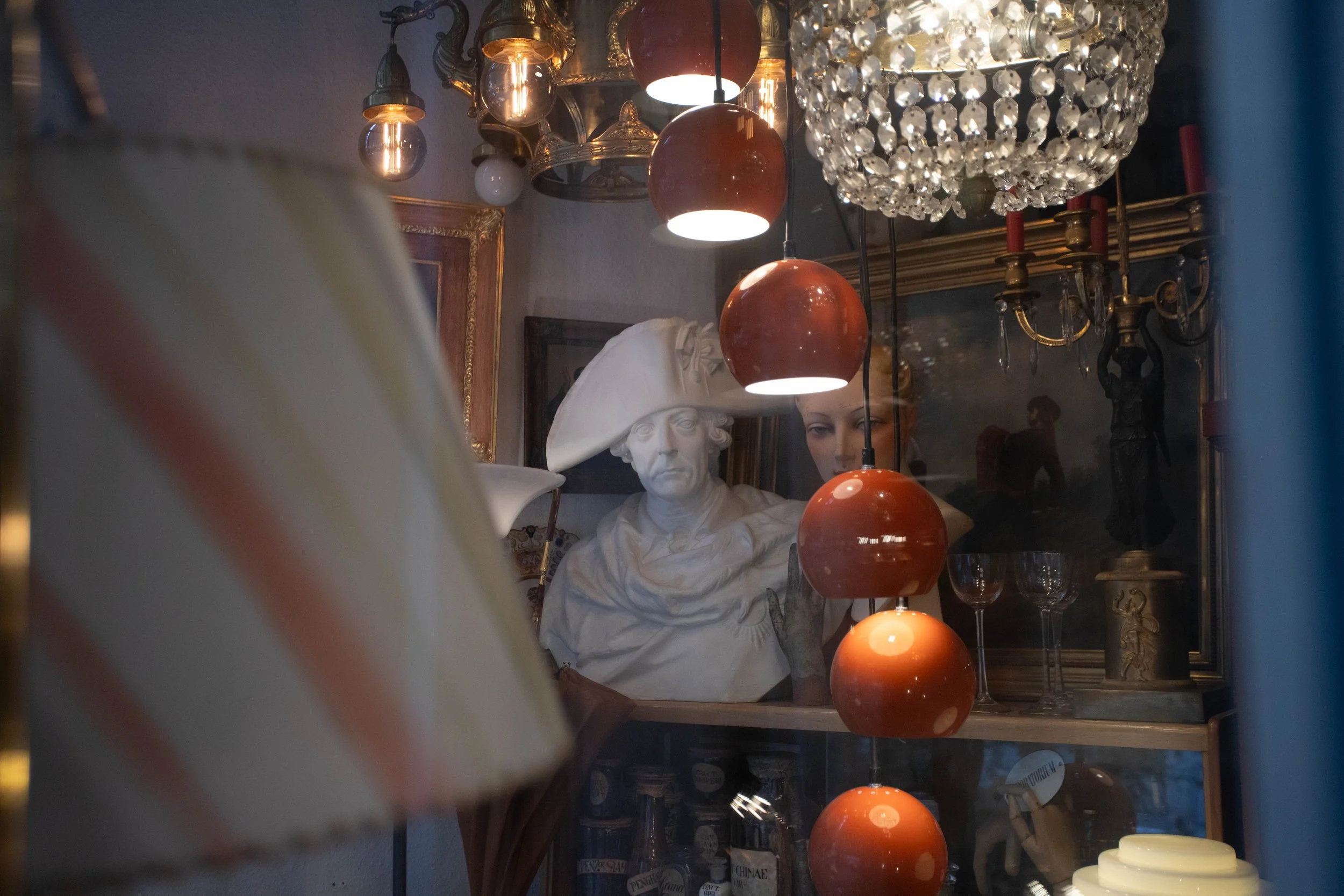 Ein Blick durch eine Ladenfensterscheibe zeigt eine Sammlung von klassischen und modernen Dekorationsobjekten, darunter eine weiße Skulptur einer Frau mit Hut, einen Kronleuchter, einen Lampenschirm, mehrere Kristalllüssel und Flaschen, sowie andere dekorative Gegenstände.