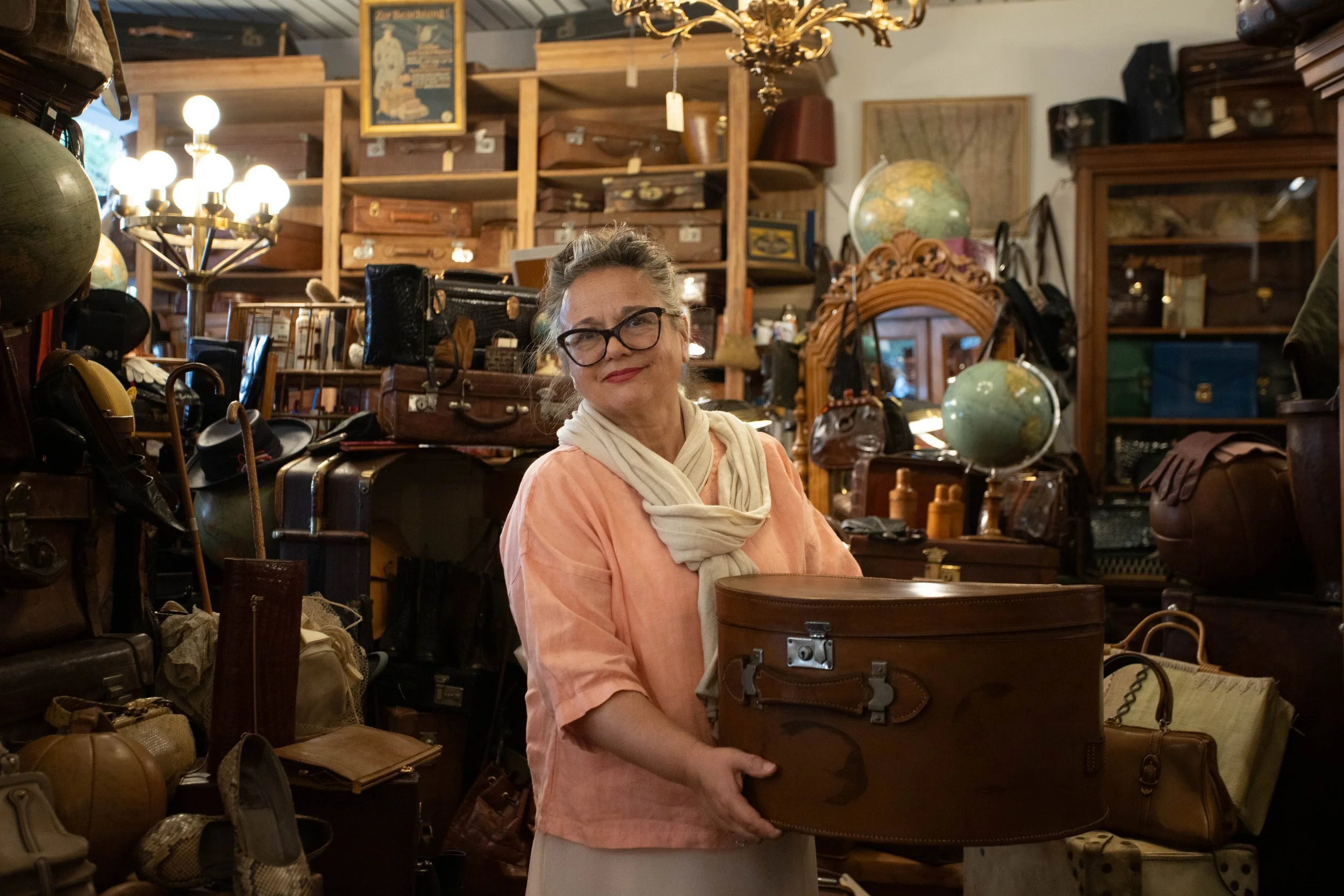 Eine ältere Frau mit Brille, weißem Schal und einem hellrosa Oberteil hält eine runde alte Tasche in einem Antiquitätenladen.