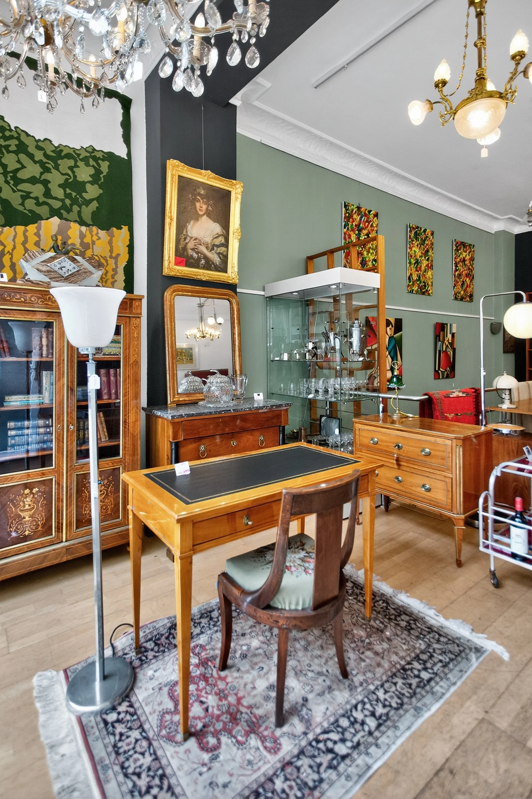 Vintage Möbel und Dekorationsgegenstände in einem Antiquitätenladen, inklusive Holzschreibtisch, Sessel, Glasvitrine, Gemälde, Spiegel, Laminatboden und Kronleuchter.