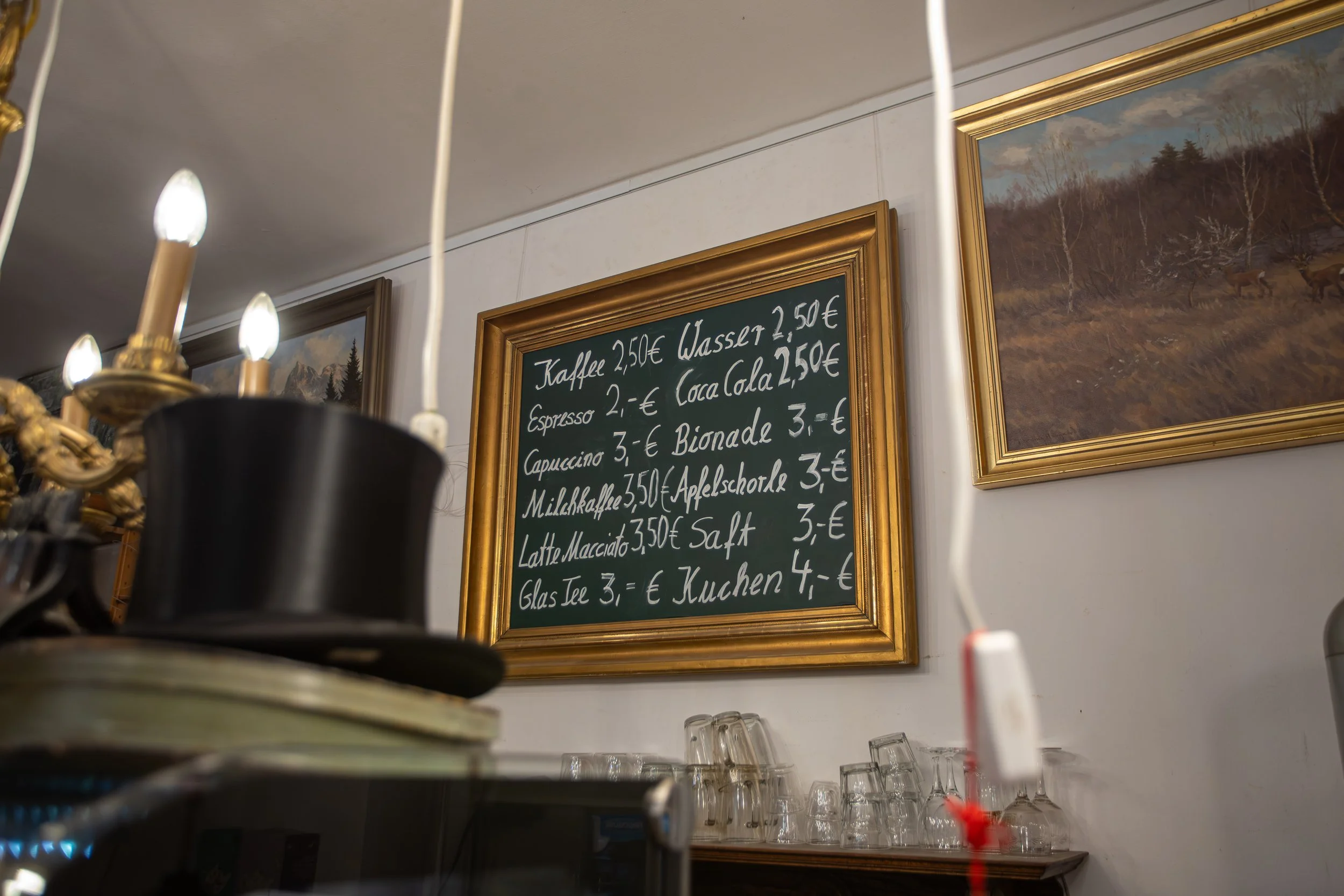 Menütafel mit Getränkepreisen in deutscher Sprache in einem Café, umgeben von gerahmten Landschaftsbildern.