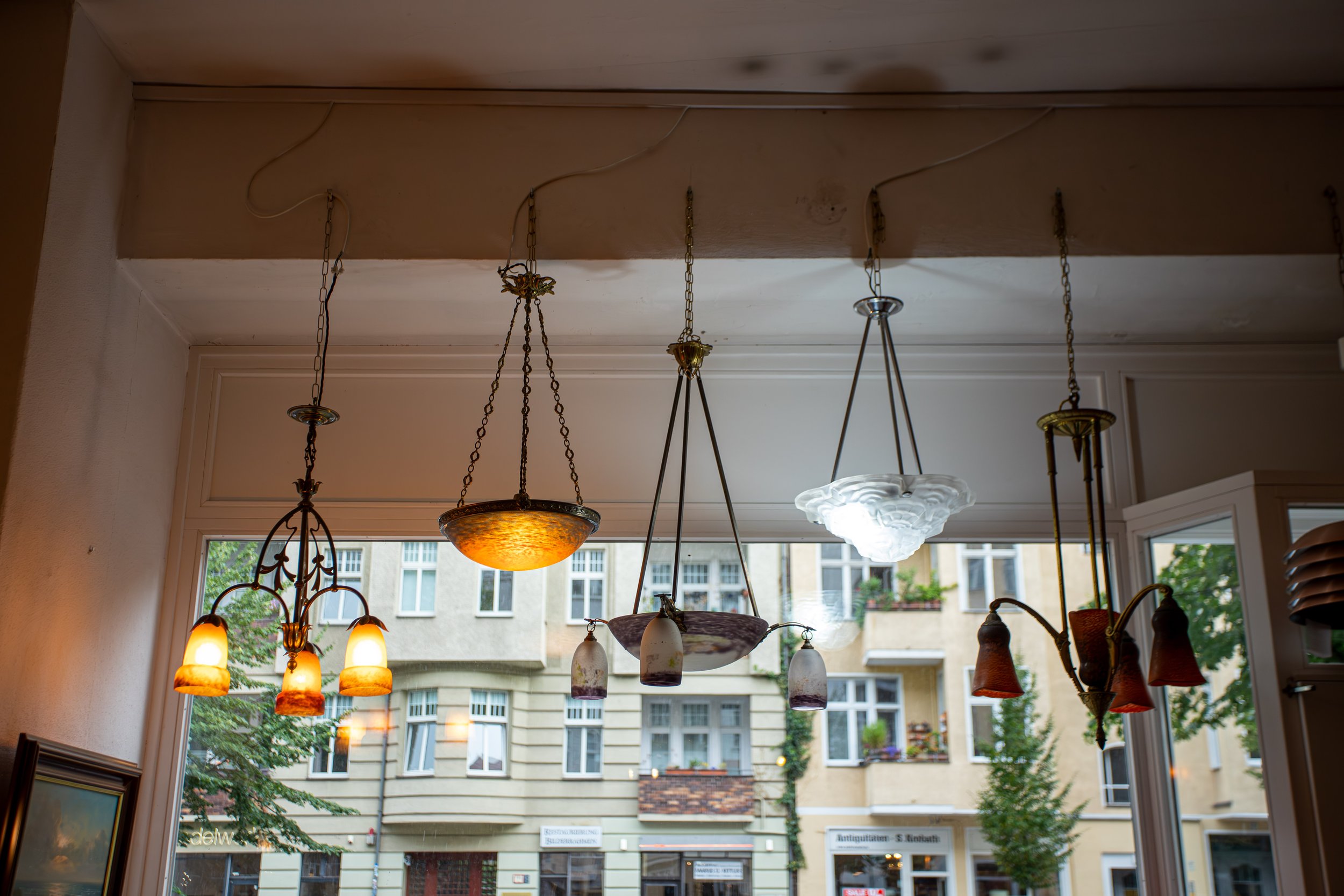 Mehrere alteingesessene Hängelampen mit verschiedenen Designs und Farben, hängen an der Decke eines Geschäfts oder einer Villa, mit Blick auf die Straße und ältere Gebäude im Hintergrund.