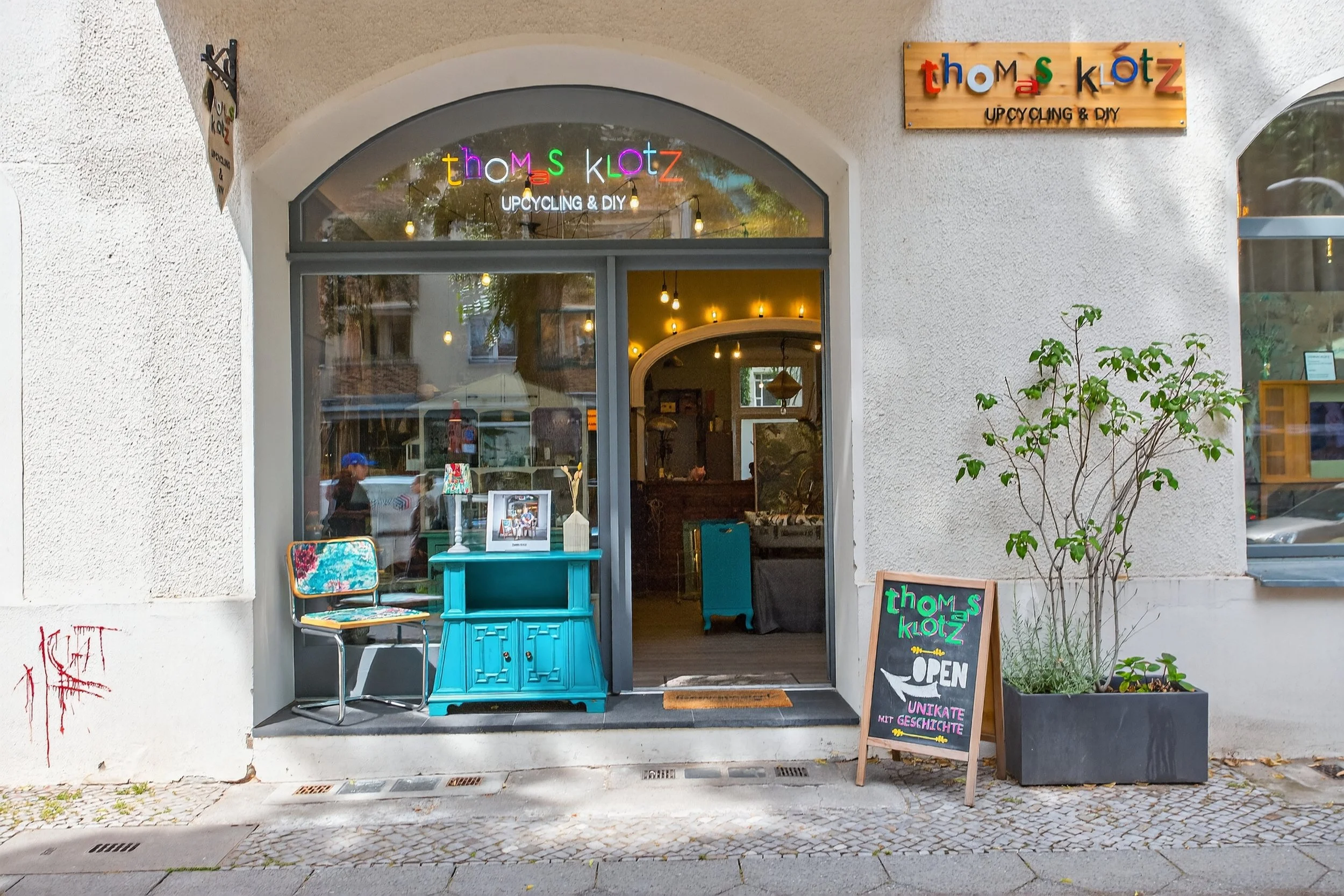 Der Eingang eines Geschäfts namens „thoms klotz“, das Upcycling und DIY anbietet. Der Laden hat eine große Fensterfront mit bunten Buchstaben am oberen Rand, die den Namen des Ladens anzeigen. Draußen vor dem Eingang steht ein schwarzes Sandwichboard mit bunten Kreidebuchstaben, das „OPEN“ und andere Hinweise zeigt. Es gibt auch eine kleine grüne Pflanzenecke neben dem Eingang.