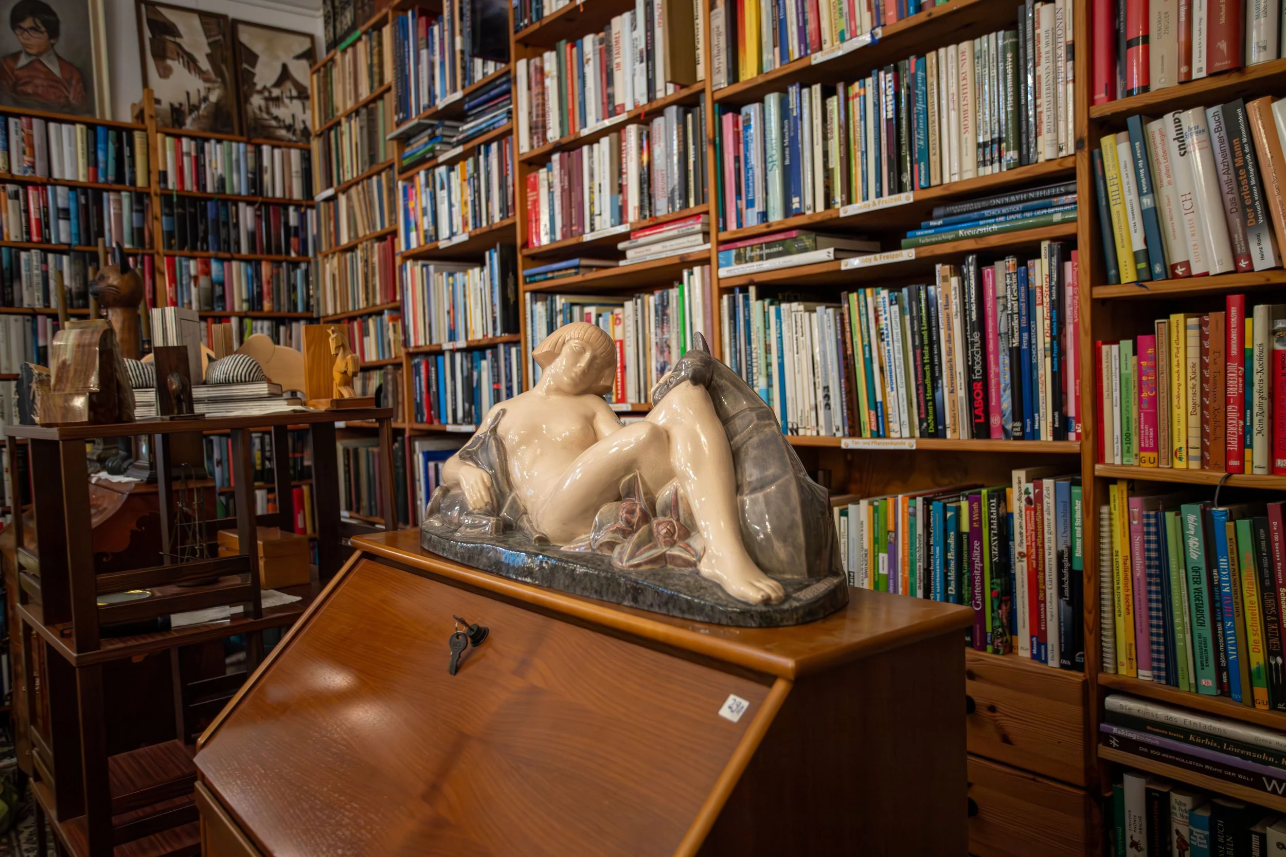 Regale voller Bücher in einer Bibliothek, darunter eine Skulptur eines liegenden nackten Mädchens auf einem Holztisch.
