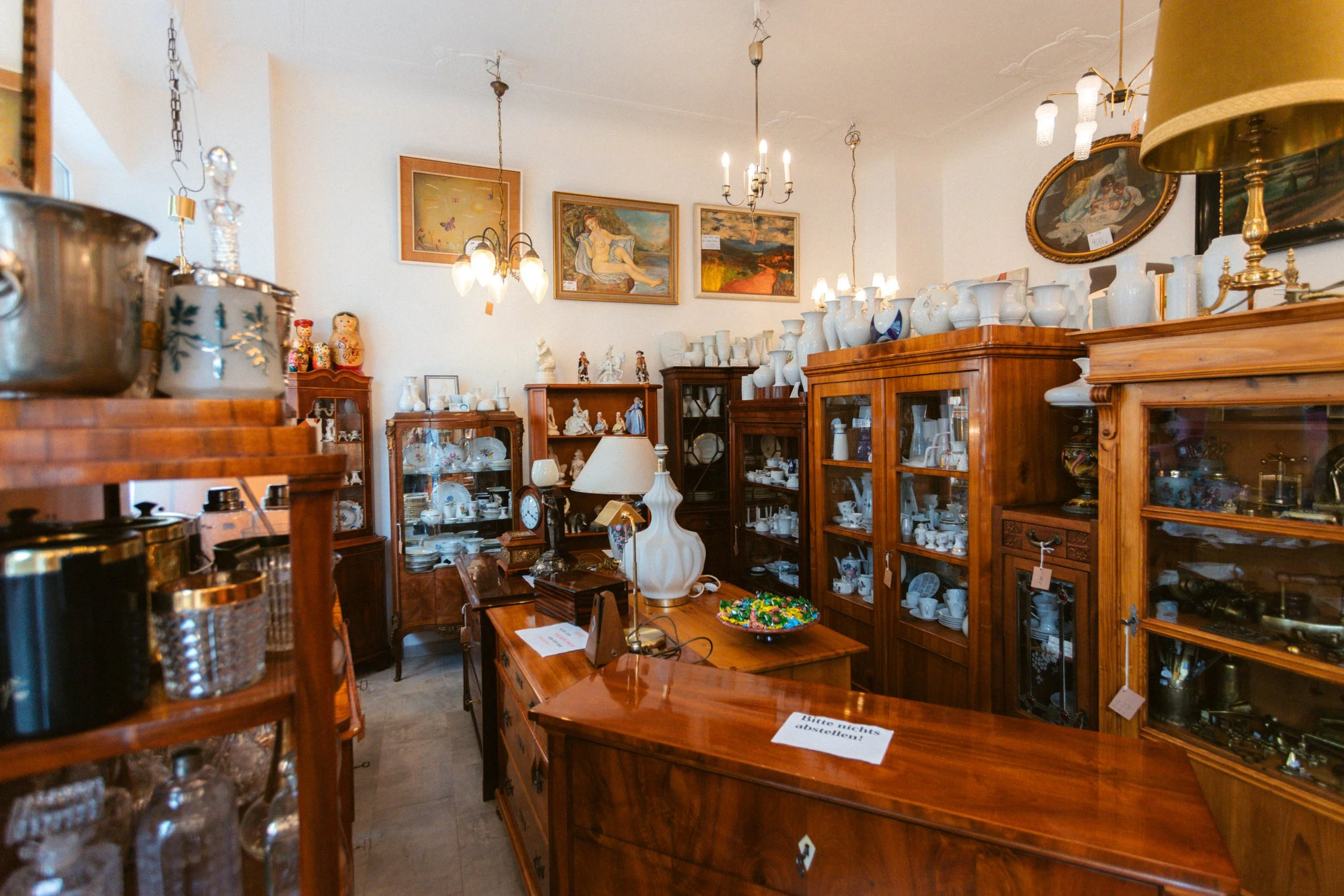 Ein Raum voll mit alten Holz- und Glaskeramik-Vitrinen, die Antik- und Sammlergegenstände wie Geschirr, Vasen und Dekorationsartikel enthalten. An den Wänden hängen Gemälde und manche Gegenstände stehen auf Tischen oder Regalen.