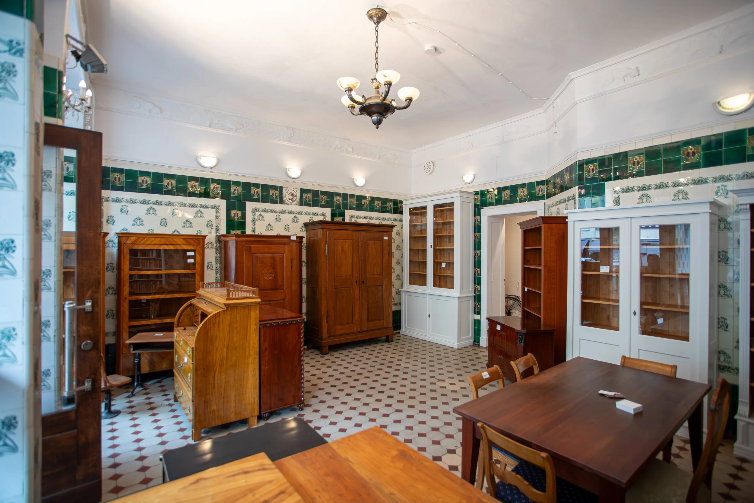 Gemütlicher Raum mit antiken Holzmöbeln, weißen und grünen Wandfliesen, Eichentischen und Stühlen, an Decke und Wänden mit klassischen Leuchten.