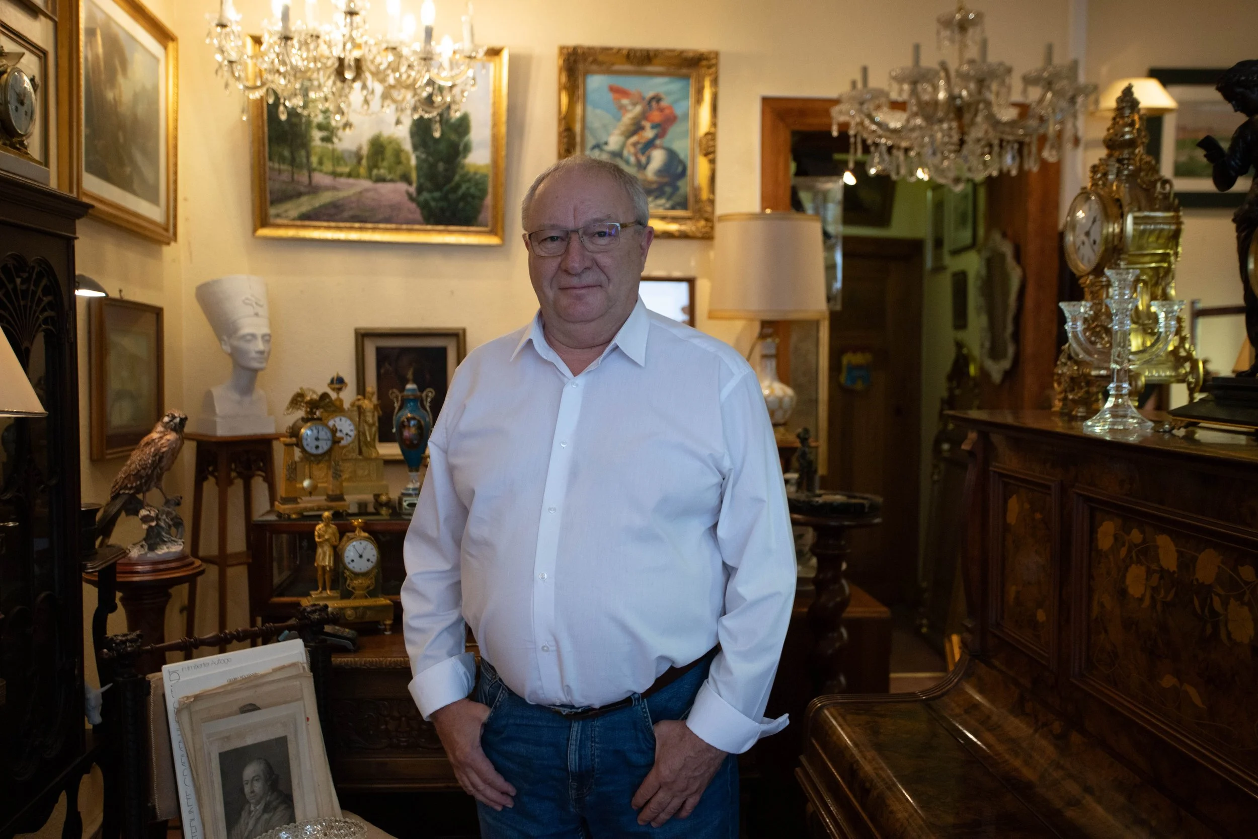 Ein älterer Mann mit Brille, weißem Hemd und Jeans steht in einem Raum voller eleganter antiker Möbel, goldener Uhren und Gemälde an der Wand.