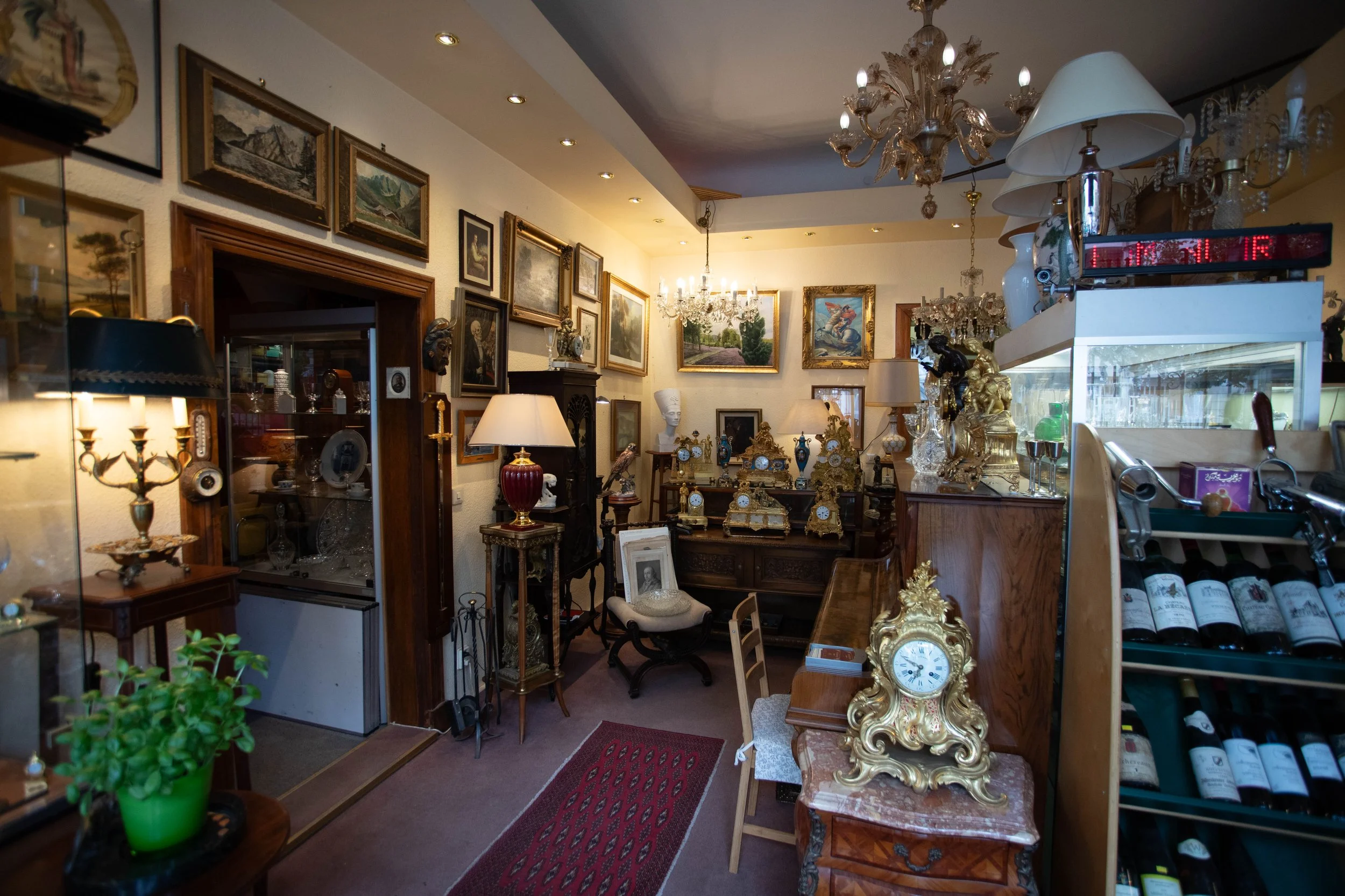 Ein Antiquitätengeschäft mit zahlreichen Gemälden an den Wänden, antiken Uhren, Vasen, Lampen und Skulpturen, sowie Weinflaschen auf einem Regal.