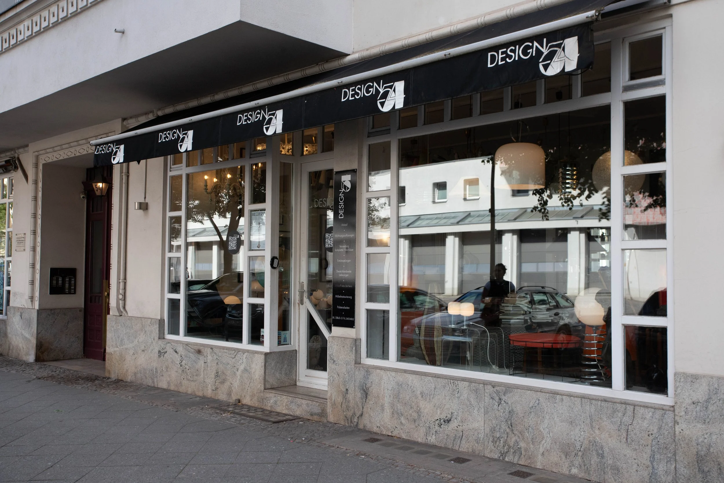 Schaufenster eines Designstudios mit großen Fenstern, Schwarzweiß-Beschriftung 'Design', Innen moderne Möbel und Lampen sichtbar, reflektierende Glasflächen und vorbeigehende Passanten.