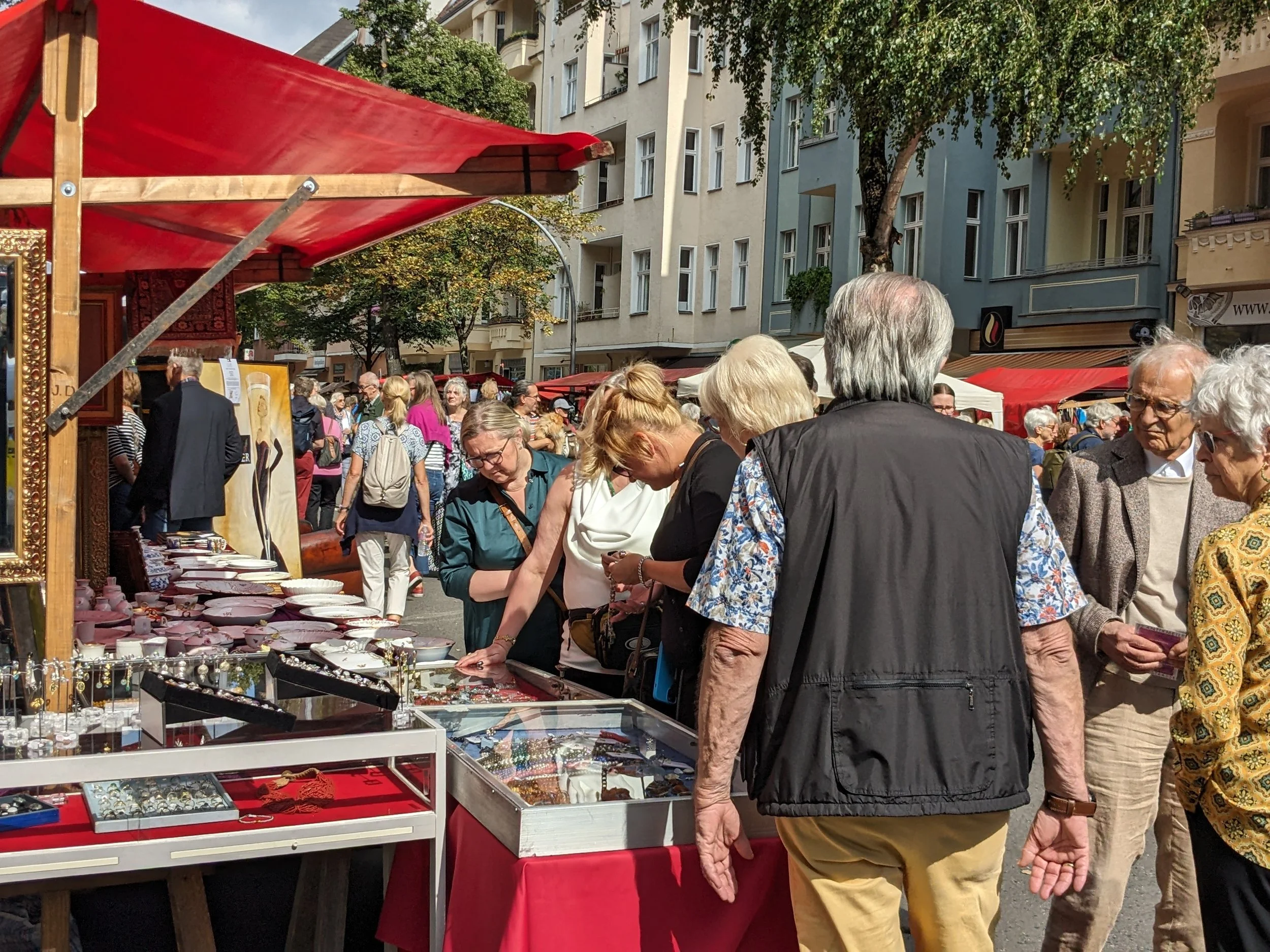 Menschen kaufen Schmuck und Accessoires an einem Straßenmarkt auf einer Sonnentag in einer Stadt. Es gibt Stände mit Kleidung und kunsthandwerklichen Gegenständen, im Hintergrund sind bunte Gebäude zu sehen.