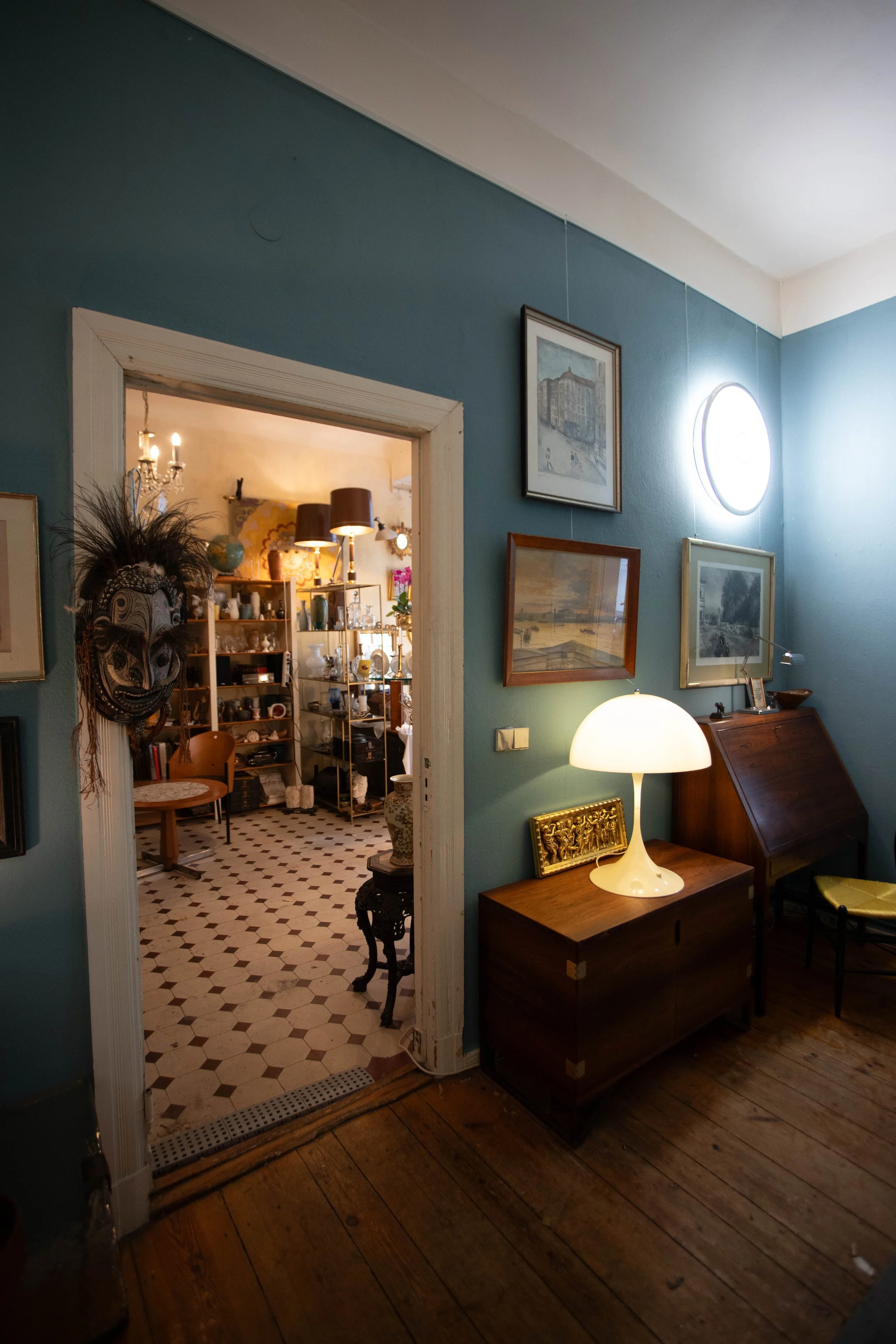 Ein Blick in ein Wohnzimmer mit blauer Wand, alten Holzfußboden und einer weißen Stehlampe auf einem Holzschrank. Im Hintergrund ein Durchgang in ein weiteres Zimmer, das mit Möbeln und Dekorationen gefüllt ist.