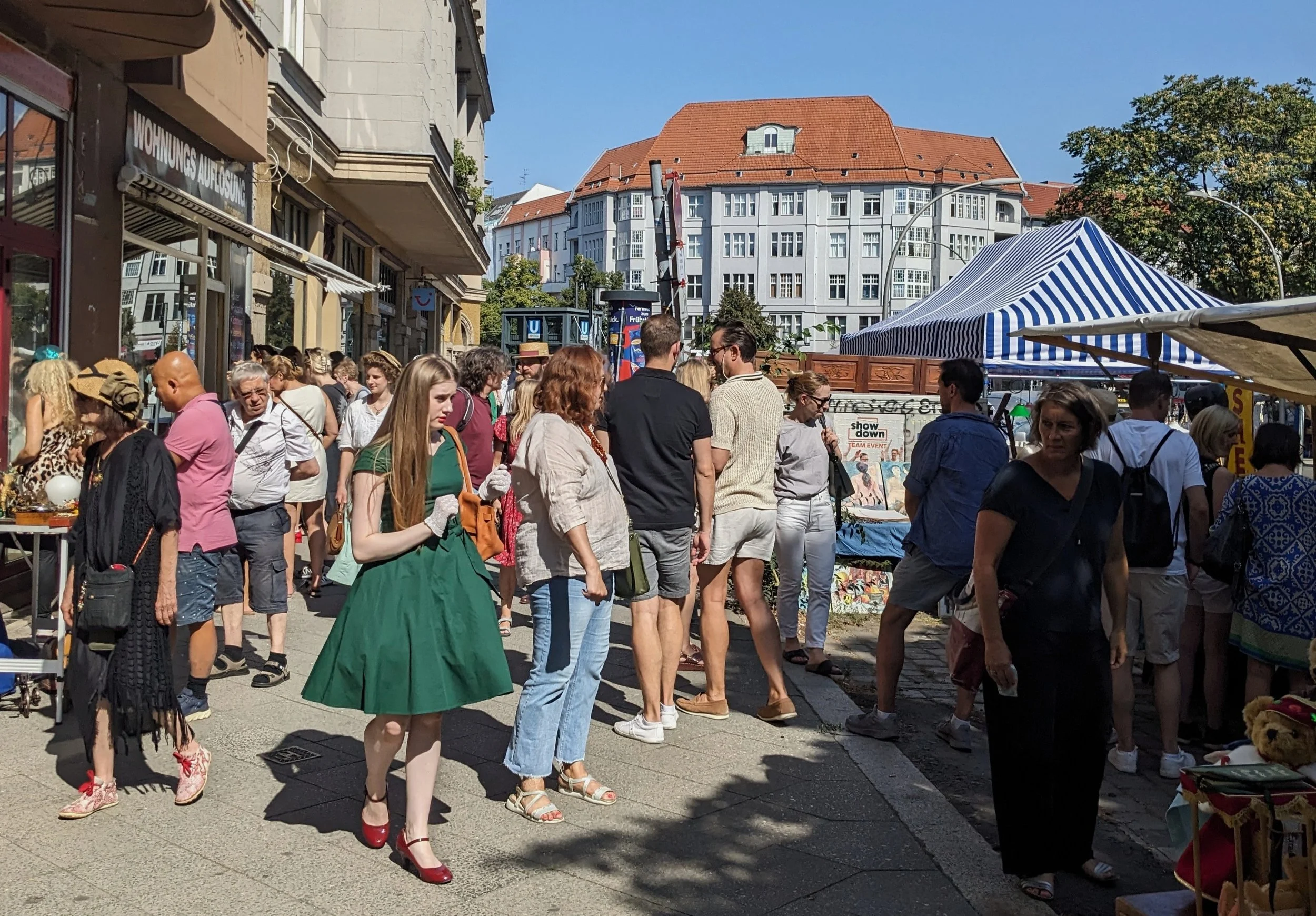 Menschen auf einem Straßenfest im Freien, mit Ständen und Zelten, am Tag mit blauem Himmel und Gebäuden im Hintergrund.