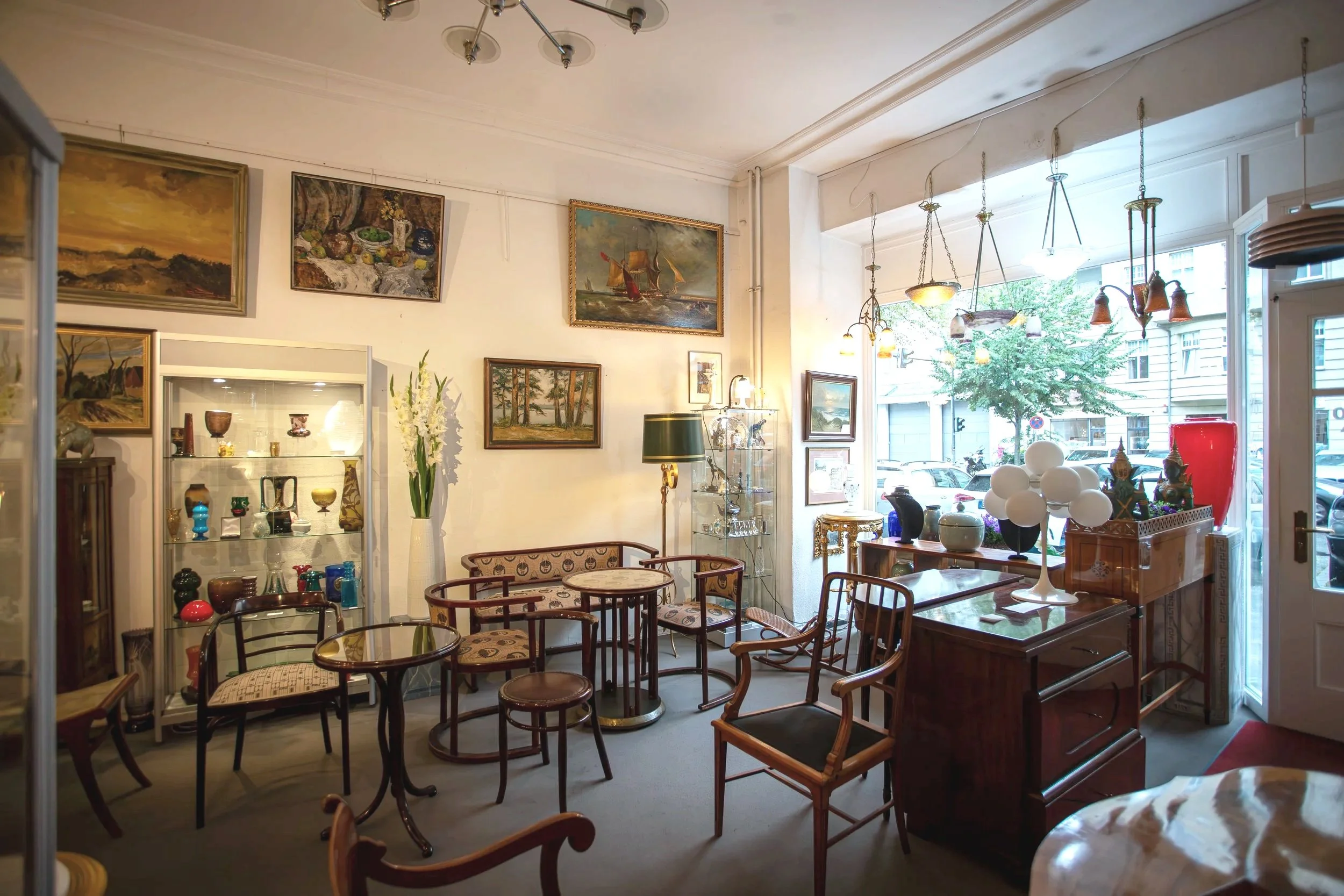 Innenraum eines antiquaren Geschäfts mit alten Möbeln, Gemälden und Dekorationsgegenständen, Blick zur Fensterfront nach draußen, mit Tischen, Stühlen und Vitrinen voller Keramiken und Skulpturen.