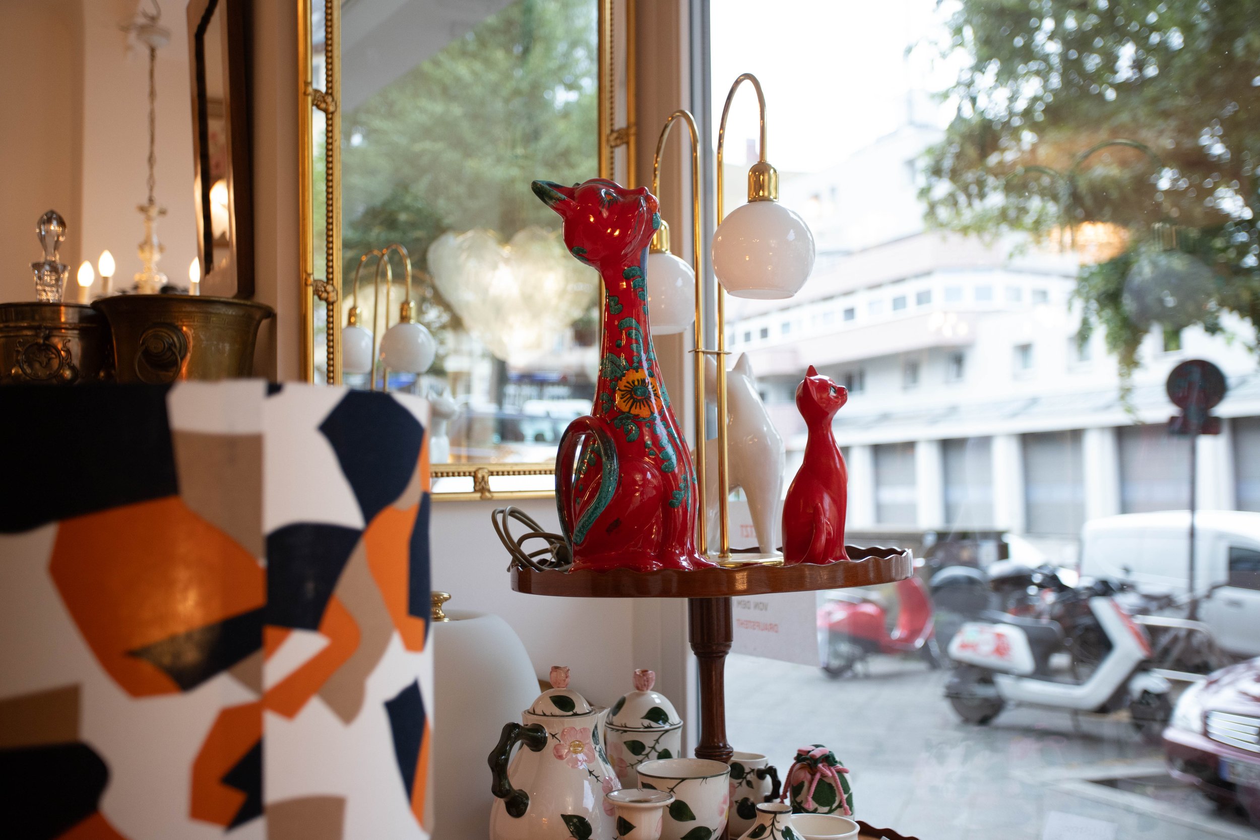 Dekorative Keramikfigurine in Form eines roten Katzen- und Hundeskulpturen, auf einem Holzständer vor einer Fensterfront mit Blick auf die Straße.