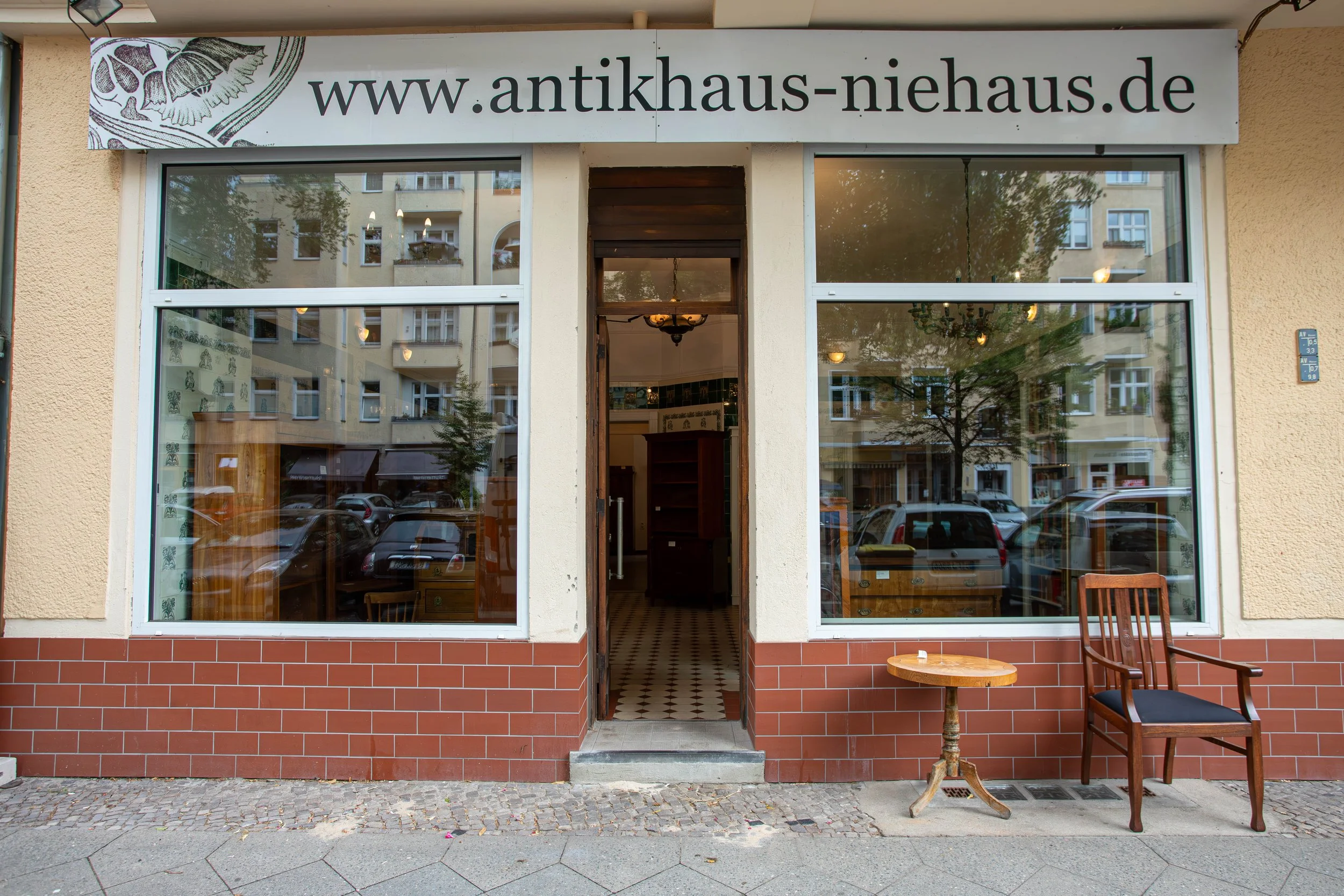 Vorgarten eines Antiquitätengeschäfts mit großen Fenstern, einem Holzsessel, einem kleinen runden Tisch, einem offenen Eingangsbereich und einem Schild mit der Webseite www.antikhause-niehaus.de.