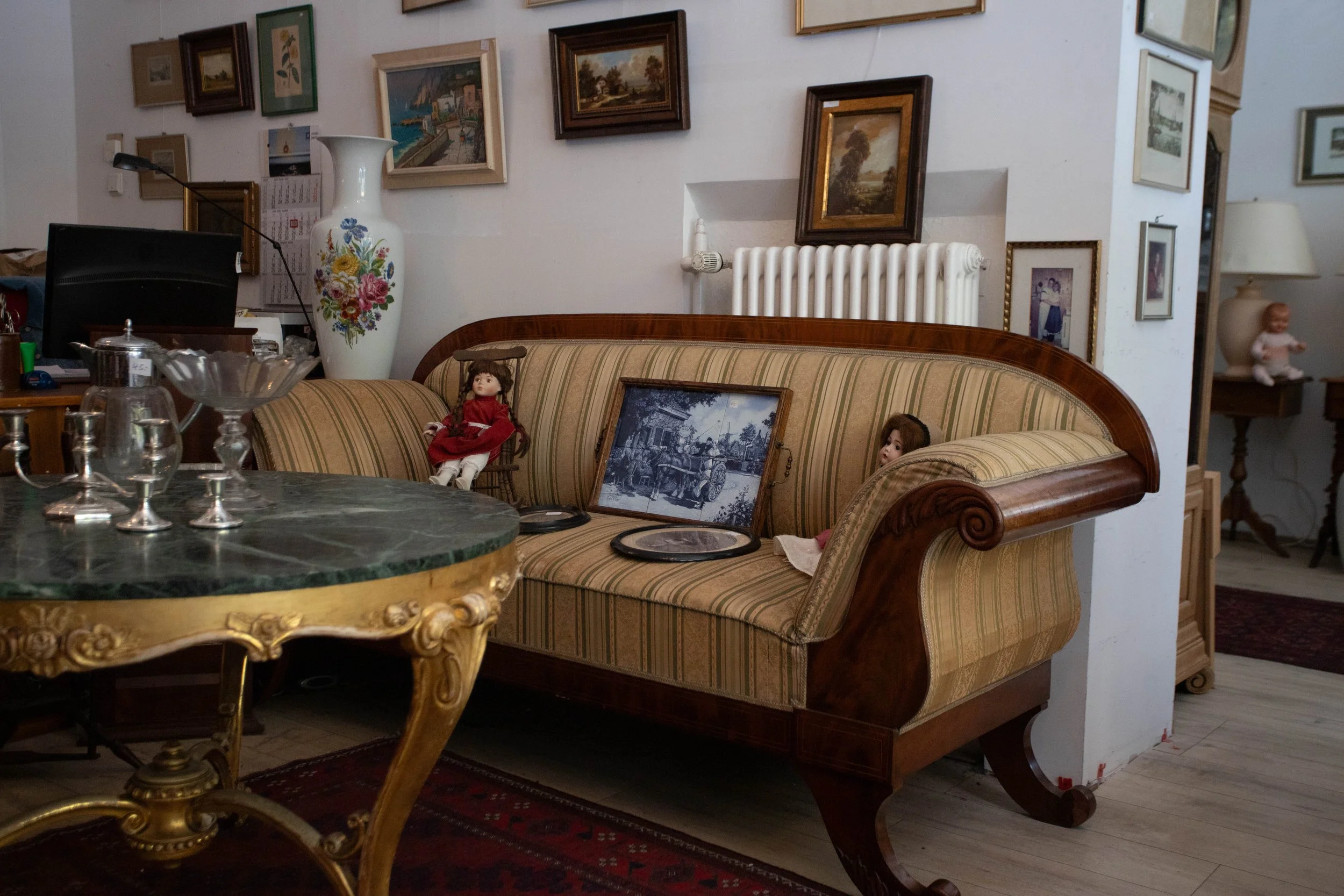 Vintage-Sofa mit gestreiftem Bezug und Holkkanten, auf einem roten Teppich, im Hintergrund Wand mit verschiedenen eingerahmten Gemälden. Auf der Couch liegen alte Porzellanpuppen und ein historisches Schwarz-Weiß-Foto.