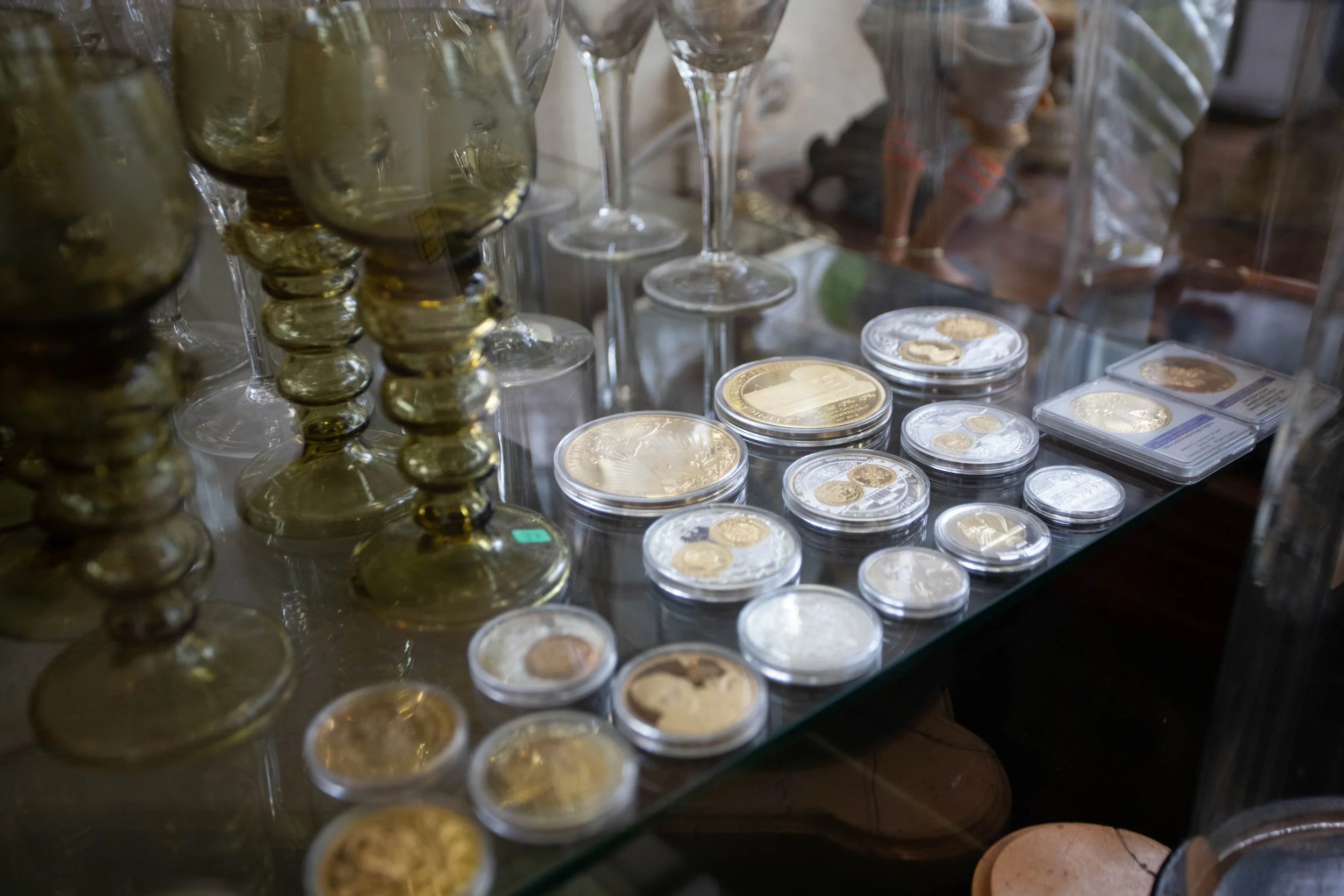 Sammlung von Münzen auf einem Glasregal, umgeben von Kerzen in goldfarbenen Halterungen und Schnitzfiguren im Hintergrund.