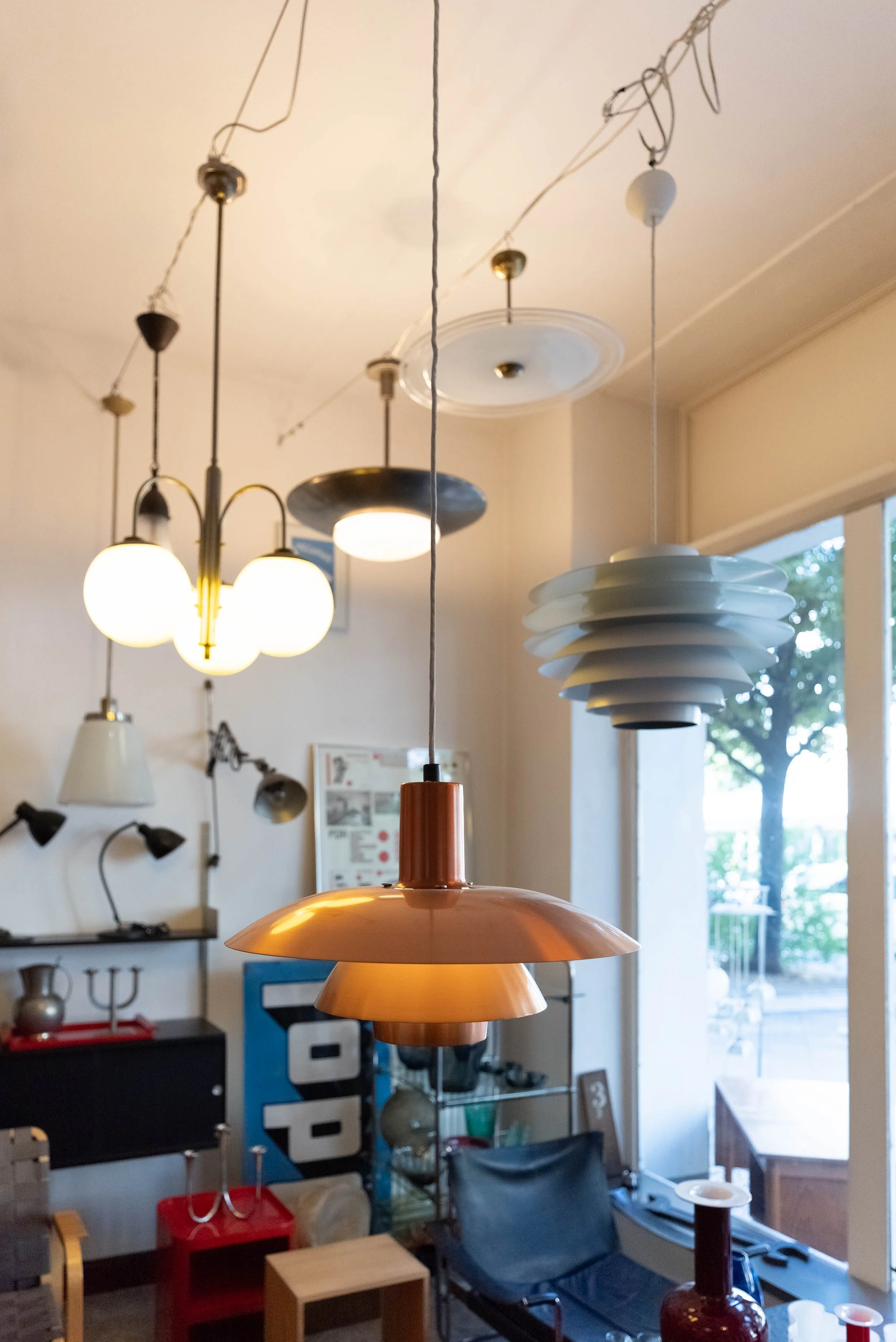 Verschiedene Designerleuchten hängen von der Decke in einem Innenraum, neben einem großen Fenster mit Blick nach draußen.