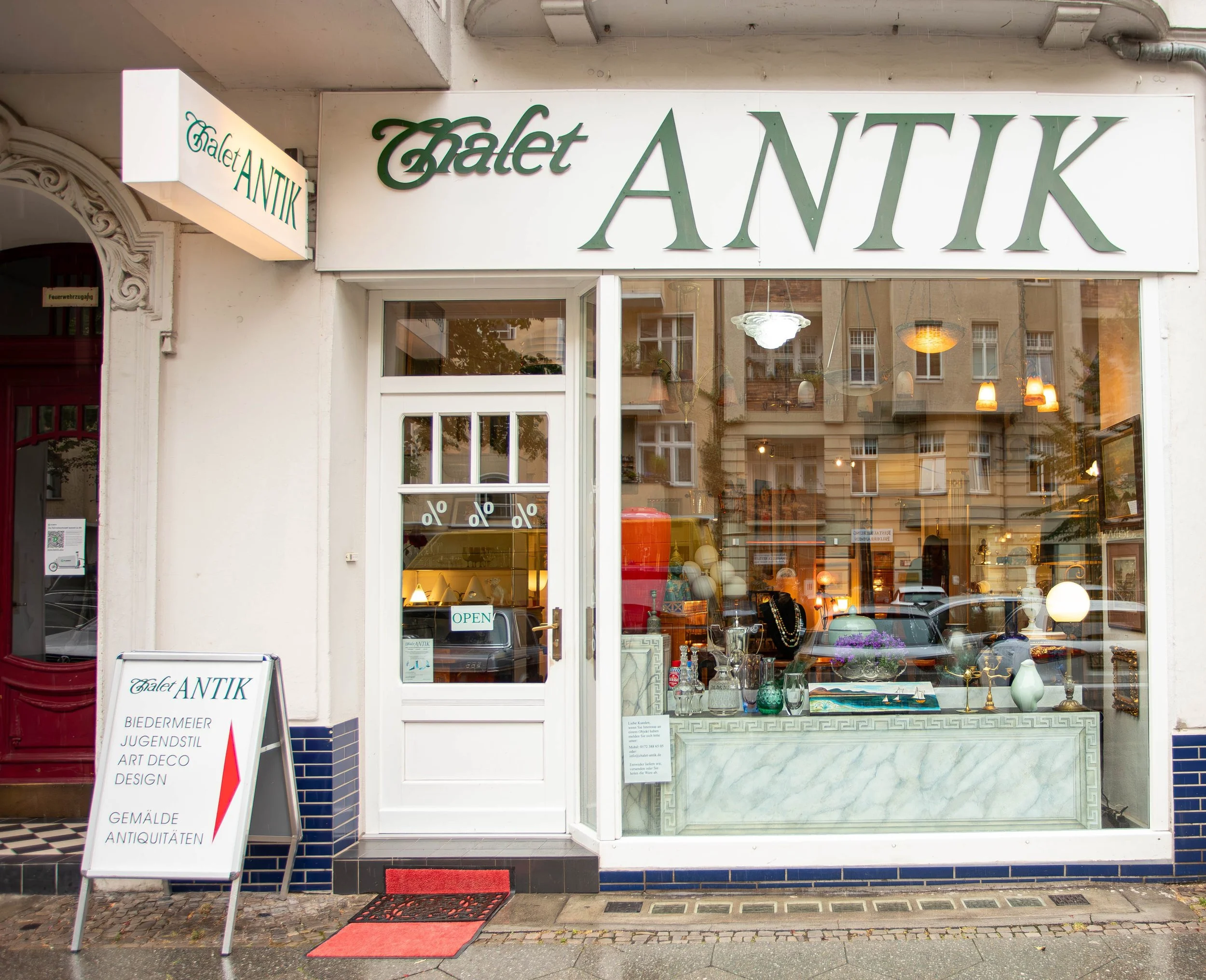 Schaufenster eines Antiquitätenladens namens 'Chalet ANTIK' mit Schmuck und Dekorationen, daneben ein Pfeil-Schild mit Adresse und Angeboten, im Hintergrund Gebäude in einer Stadtstraße.