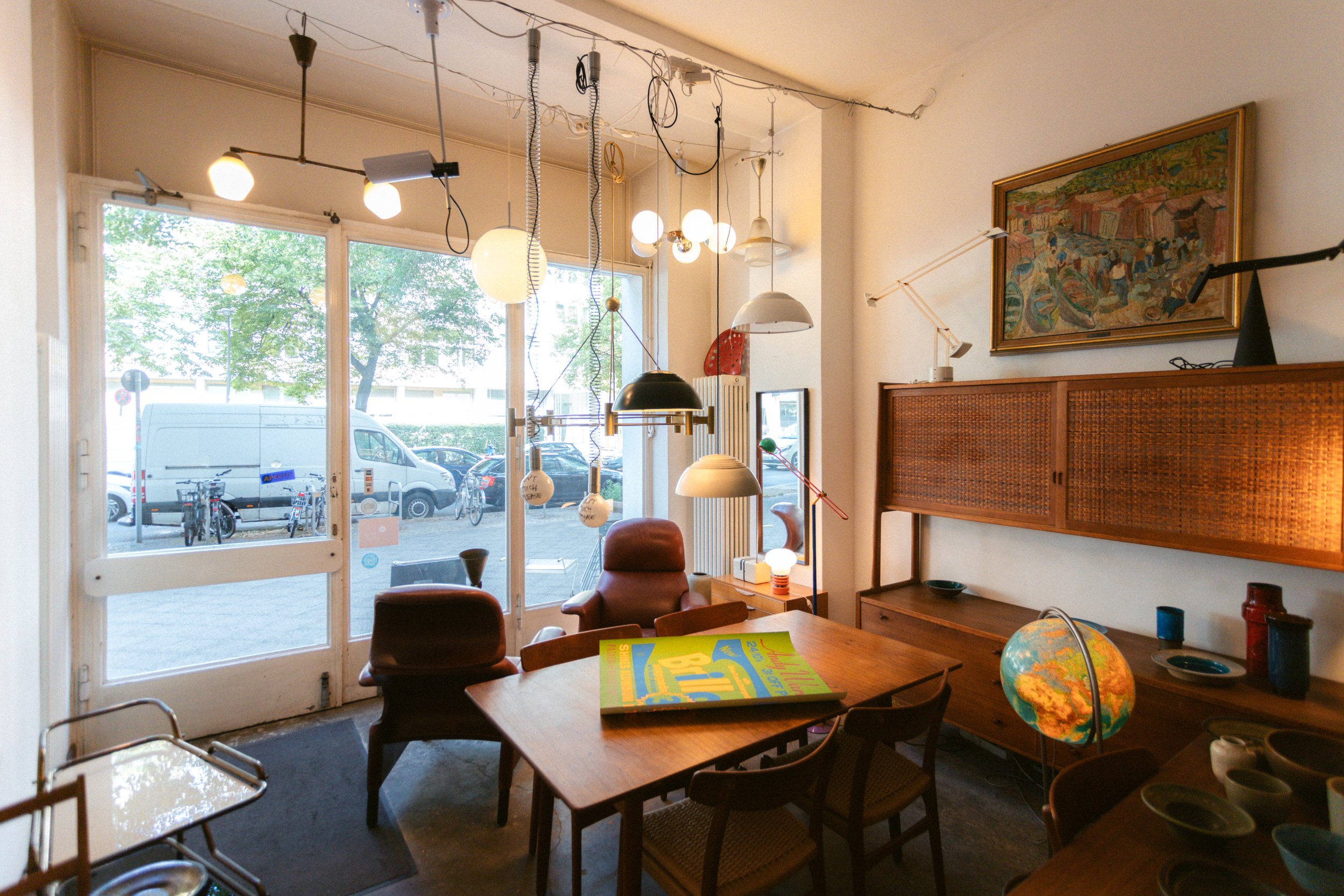 Innenraum eines gemütlichen Cafés mit Holztisch, verschiedenen Stühlen, Wanddeko und mehreren Hängelampen, große Fenster mit Blick auf eine Straße mit Fahrrädern und parkenden Autos.