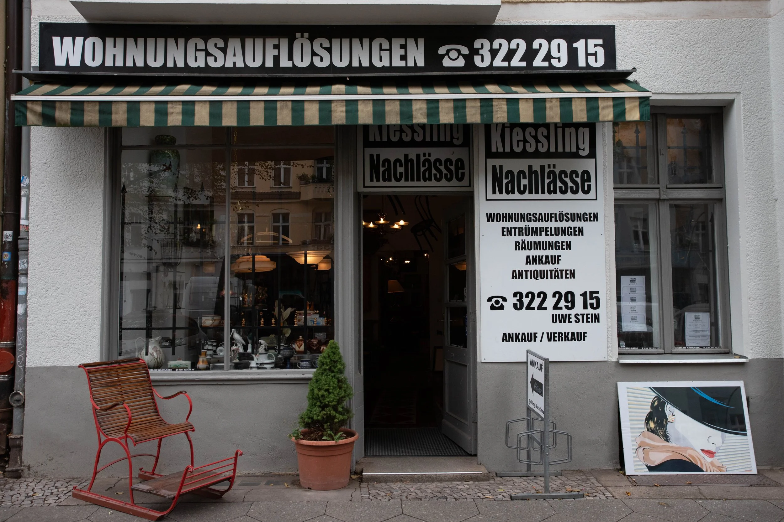 Geschäft mit Schildern, die Haus- und Wohnungsauflösungen, Entrümpelungen, Räumungen, Ankauf und Antiquitäten anbieten, mit einem roten Schlitten, einer kleinen Pflanze und einem Kunstbild im Fenster.