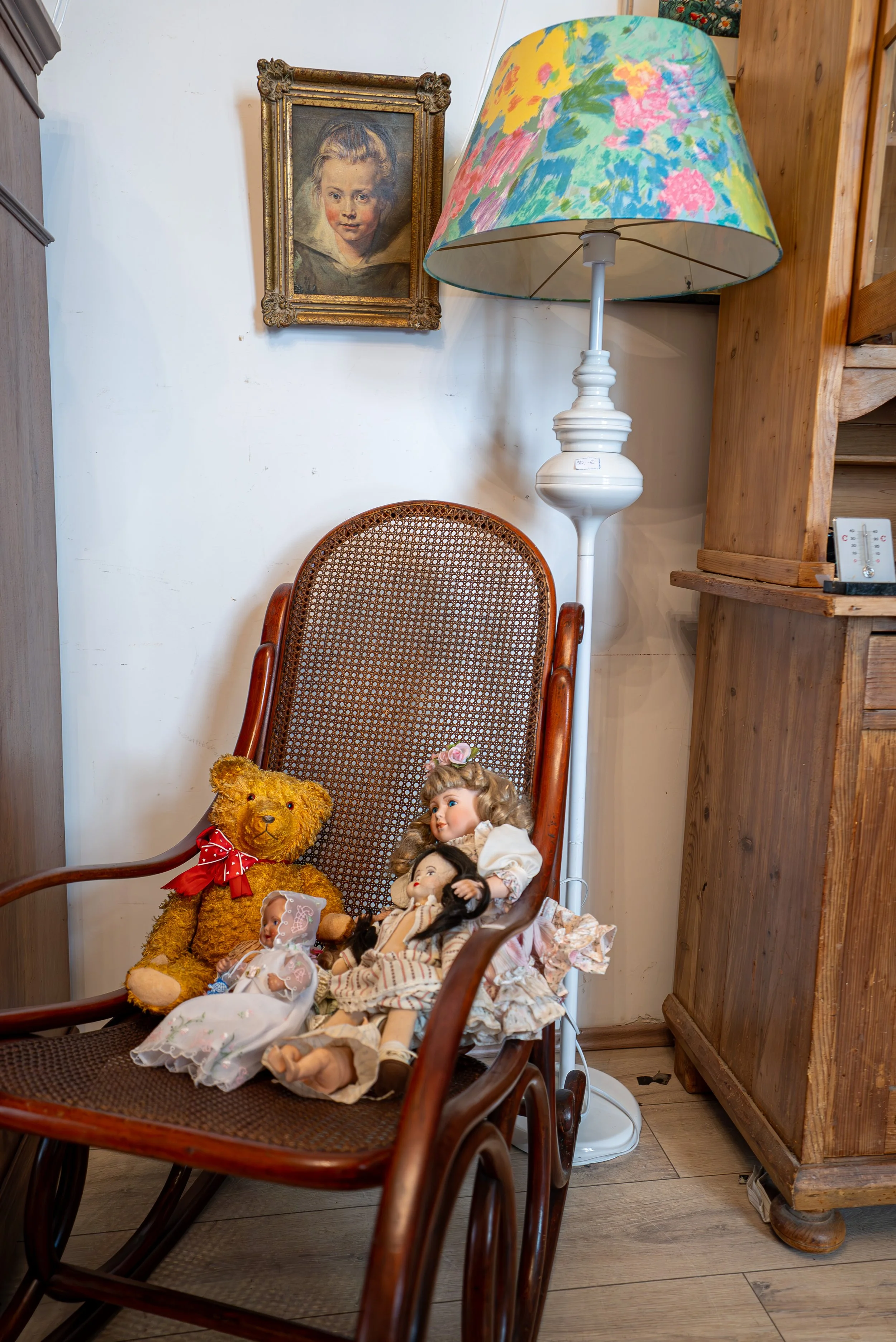 Ein Holzschaukelstuhl mit drei Puppen, einem Stoffbär mit rotem Halstuch, steht neben einem weißen Stehlampe mit buntem Lampenschirm und einem alten Gemälde eines Jungen an der Wand.