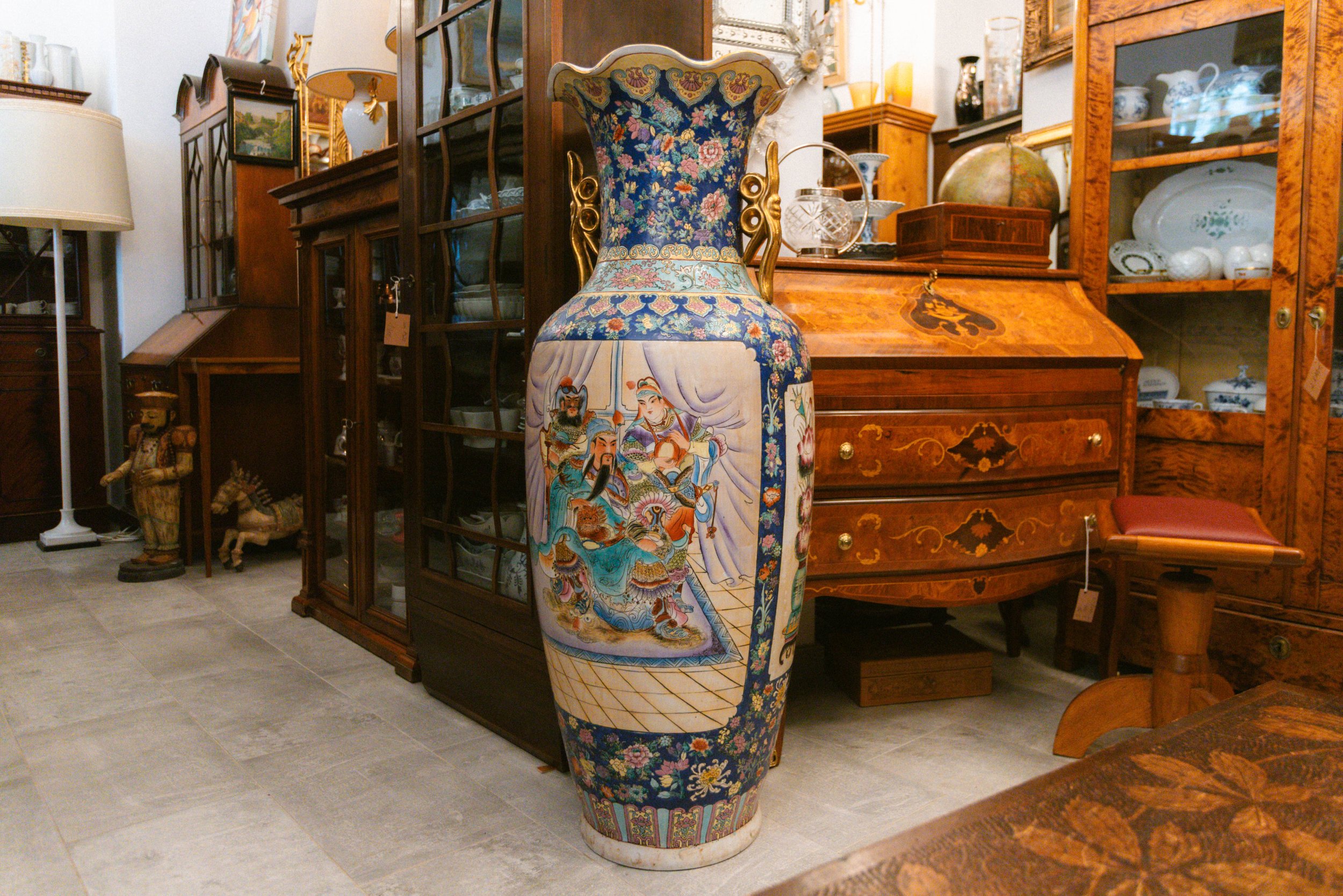Eine große, bunte asiatisch dekorierte Vase mit handgemachten Gemälden, die traditionelle Szenen zeigen. Sie steht in einem Antiquitätengeschäft neben alten Holzmöbeln und Geschirr.