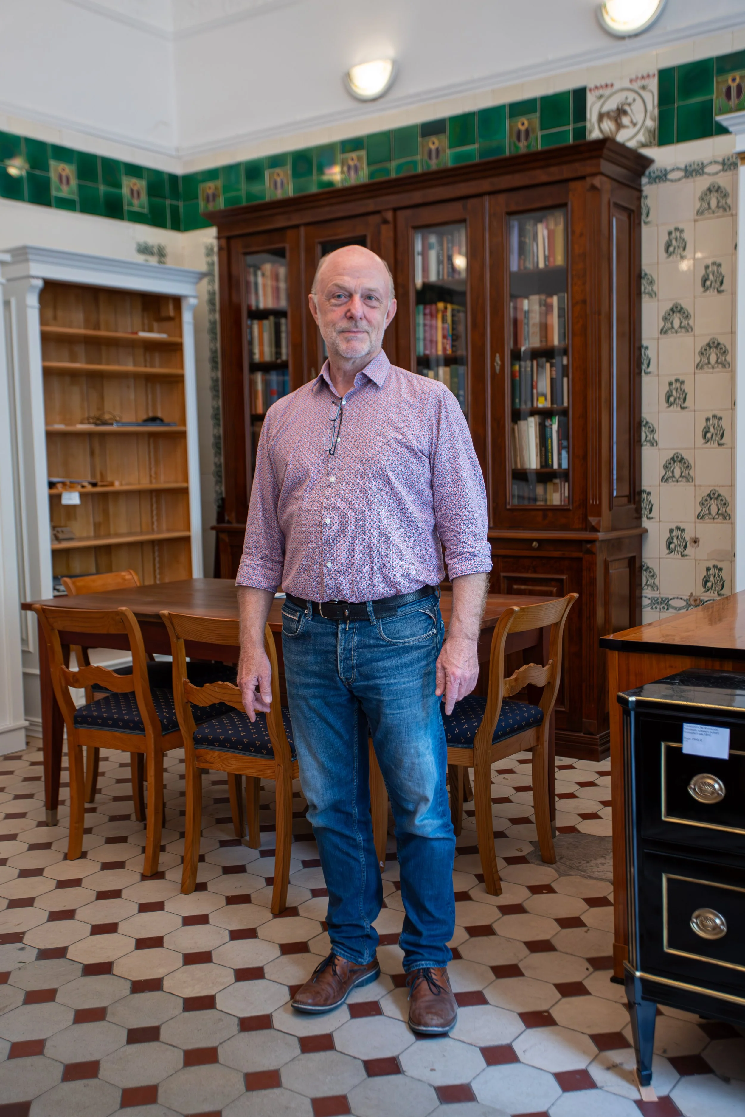 Ein älterer Mann in Hemd und Jeans steht in einem rustikalen Raum mit Holzmöbeln und Bücherregalen.