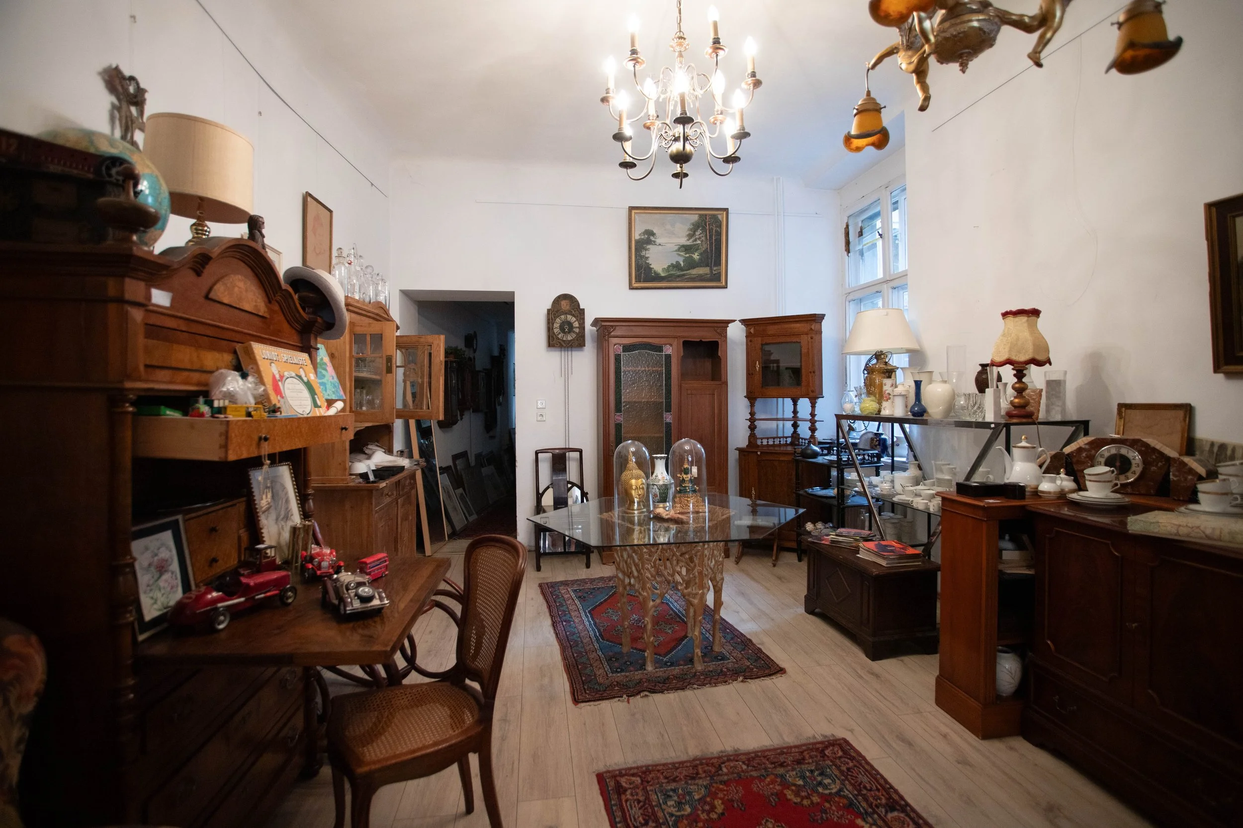 Ein Wohnzimmer mit antikem Holzmobiliar, einem Kronleuchter an der Decke, mehreren Teppichen, einem Tisch in der Mitte mit Glastischplatte und dekorativen Glasschirmen, sowie einem Fenster an der rechten Wand, das Tageslicht hineinscheinen lässt.