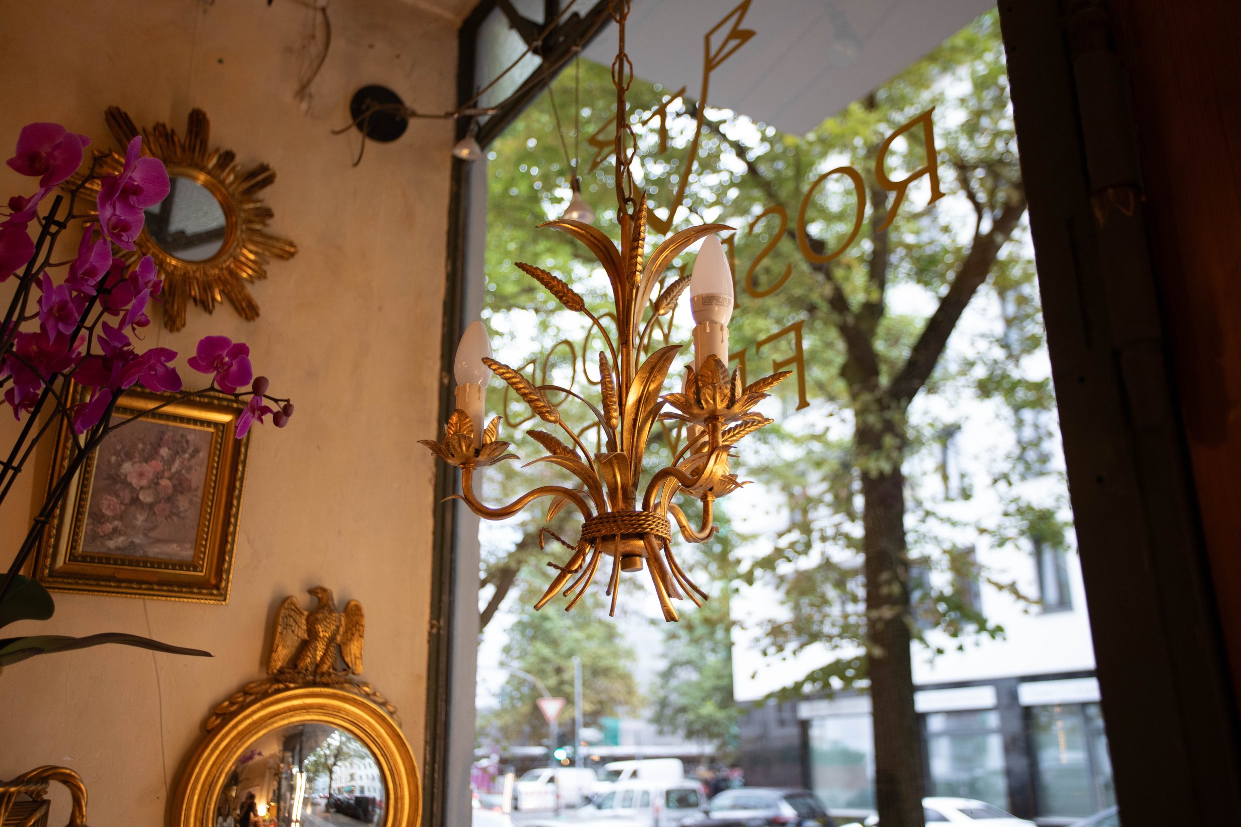 Ein goldener Kronleuchter mit zwei Lampen hängt vor einer Fensterfront mit Blick auf Bäume und Fahrzeuge auf der Straße. Im Hintergrund sind Wanddekorationen, darunter ein kleiner Spiegel und ein goldener Fasenrahmen, sichtbar.