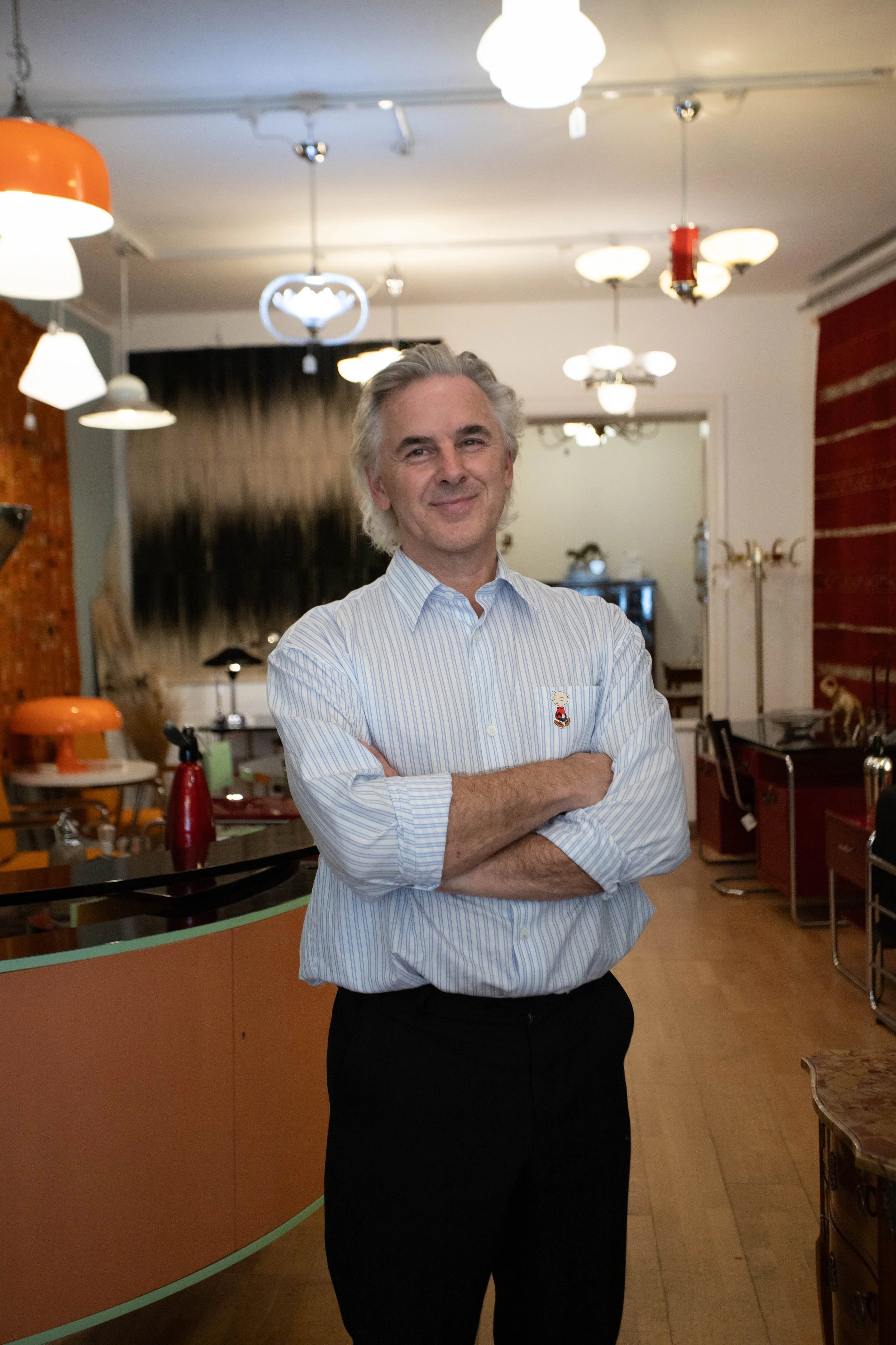 Ein Mann mit grauem Haar, in einem weißen Hemd mit blauen Streifen, steht in einem Raum mit bunter Dekoration, Lampen an der Decke und kleinen dekorativen Gegenständen im Hintergrund.