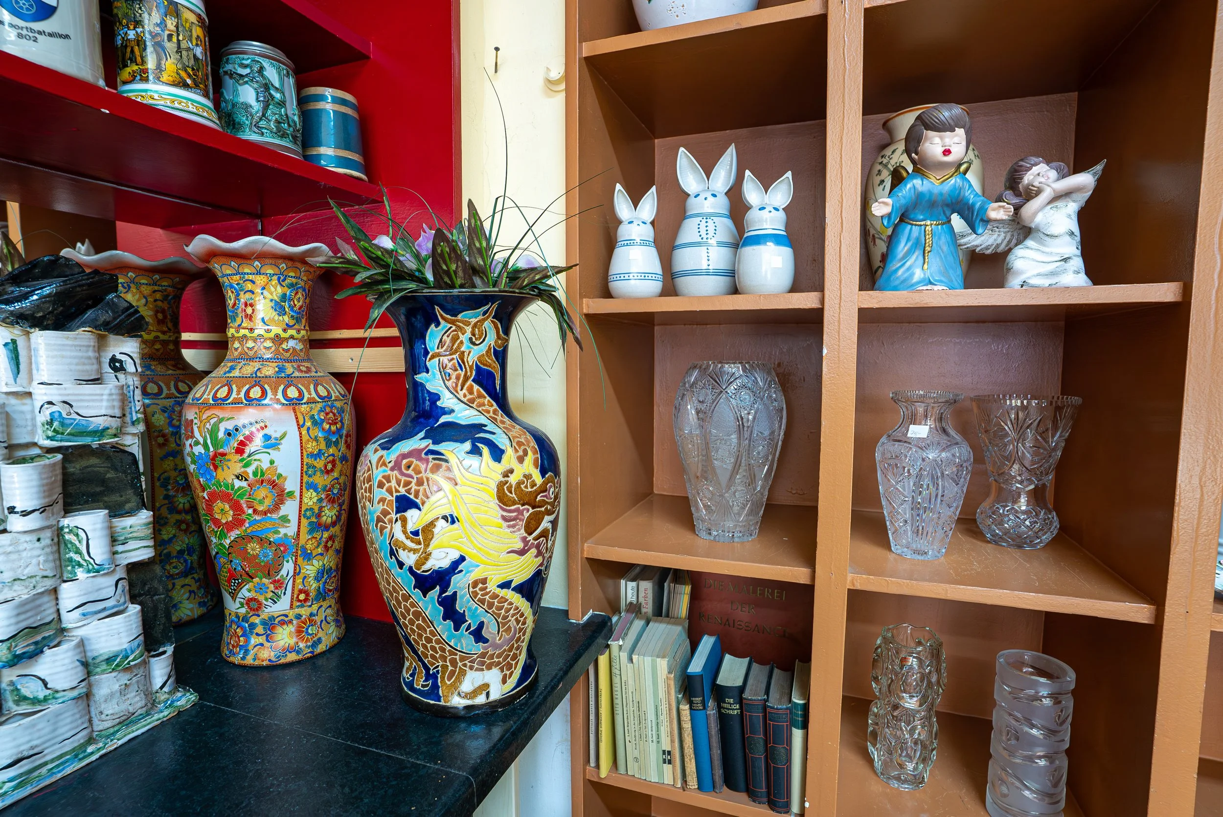 Verschiedene Keramiktöpfe, Gläser und Figuren auf Holzregalen, inklusive Mingu-Keramikvase mit Blumen, Engel-Figuren, Kristallvasen und Bücher.