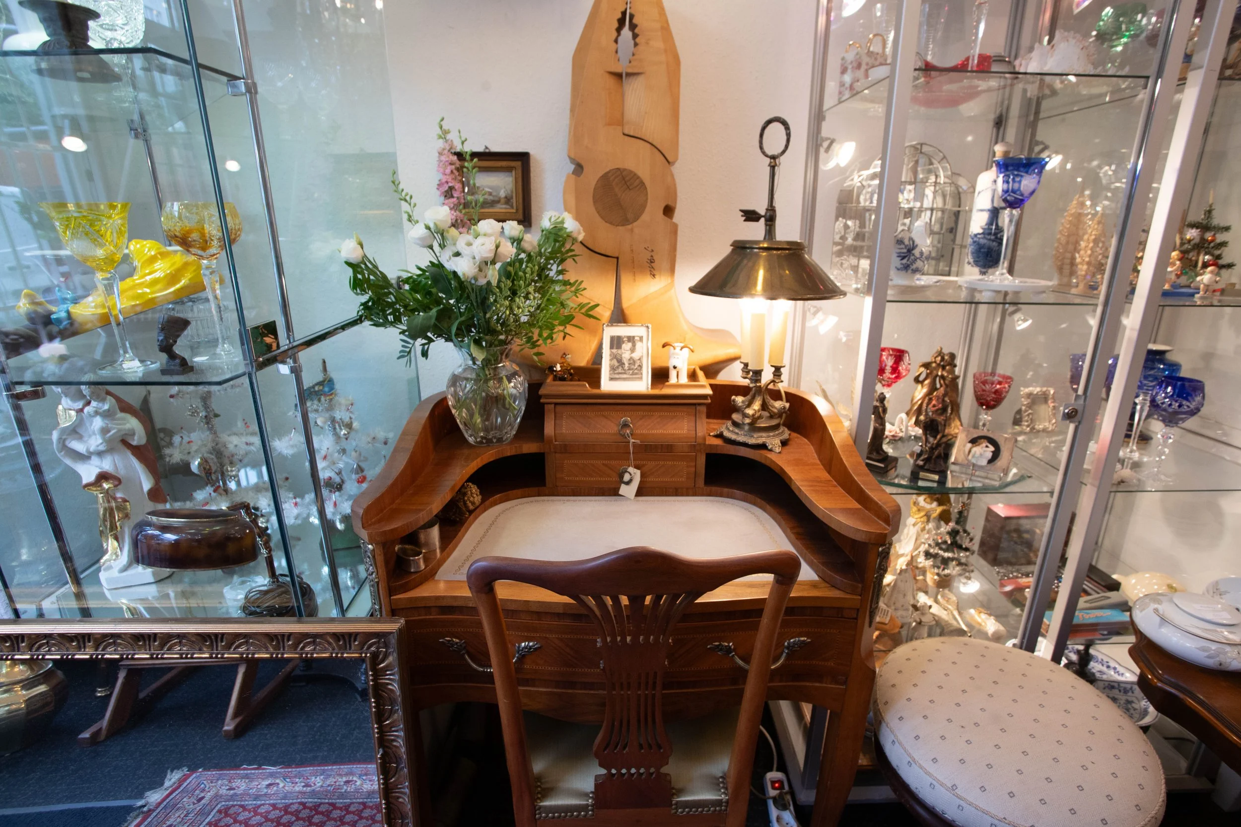 Antikes Holzschreibpult mit Blumen in Vase, daneben ein Stuhl, auf dem Pult eine Lampe, Fotografien und Dekorationsobjekte, im Hintergrund Glas-Vitrinen mit Glasvasen, Figuren und Dekorationsartikeln, in einem Raum mit Teppichboden.