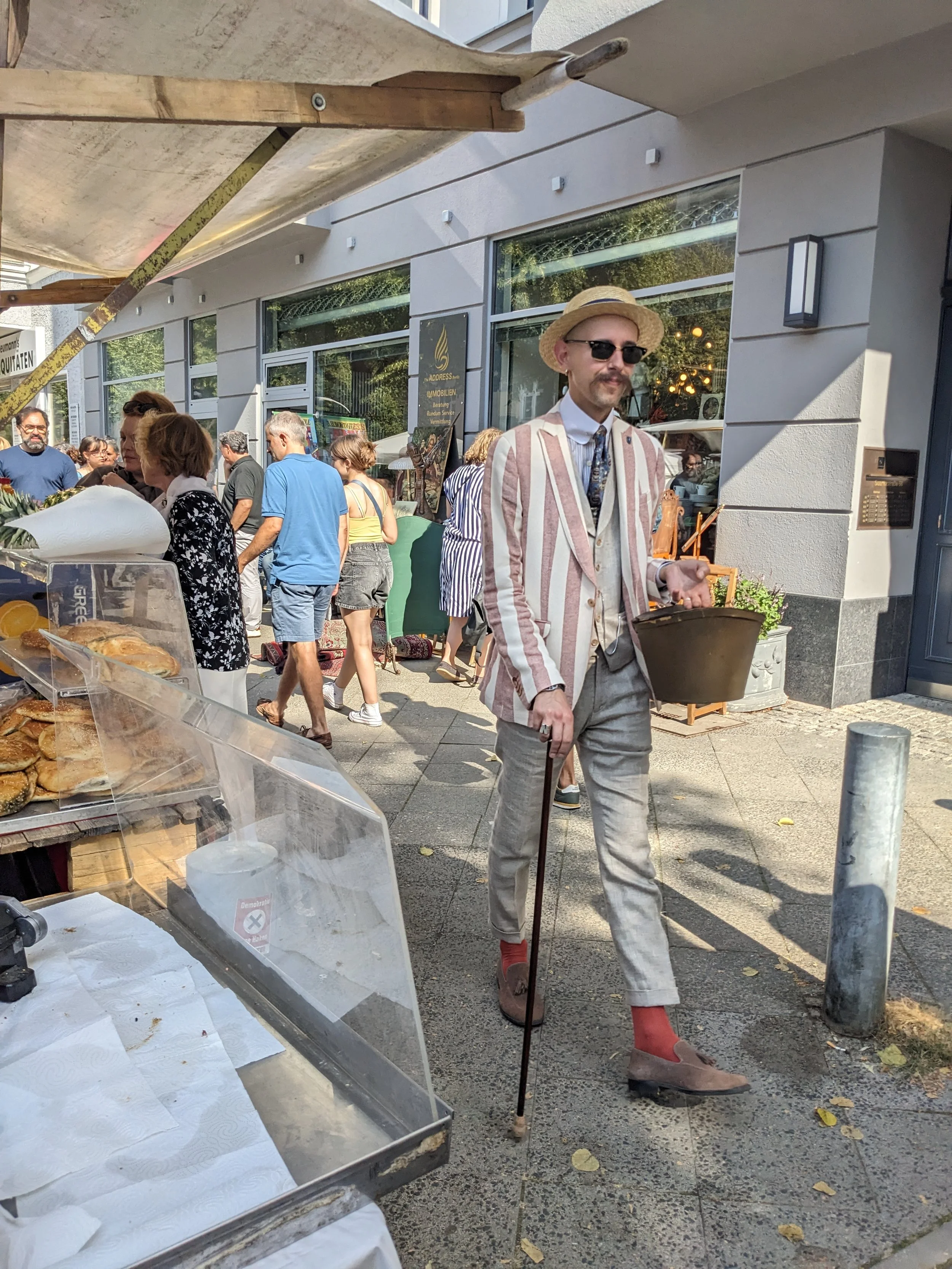 Ein Mann in einem bunten gestreiften Anzug mit Hut, Sonnenbrille und Latschen läuft auf der Straße, während um ihn herum Menschen einkaufen und sich unterhalten.