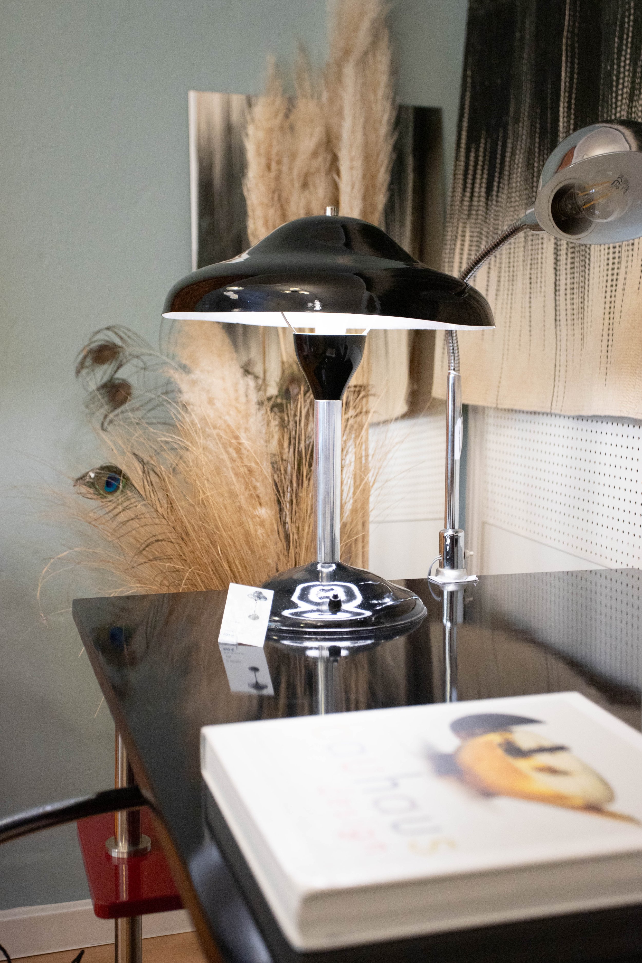 Ein schwarzer Tischlampenschirm auf einem Holzstuhl, eine Klemme mit Glühbirne, ein Buch mit einer Eistorte auf dem Cover, dekorative Trockenpflanzen im Hintergrund.