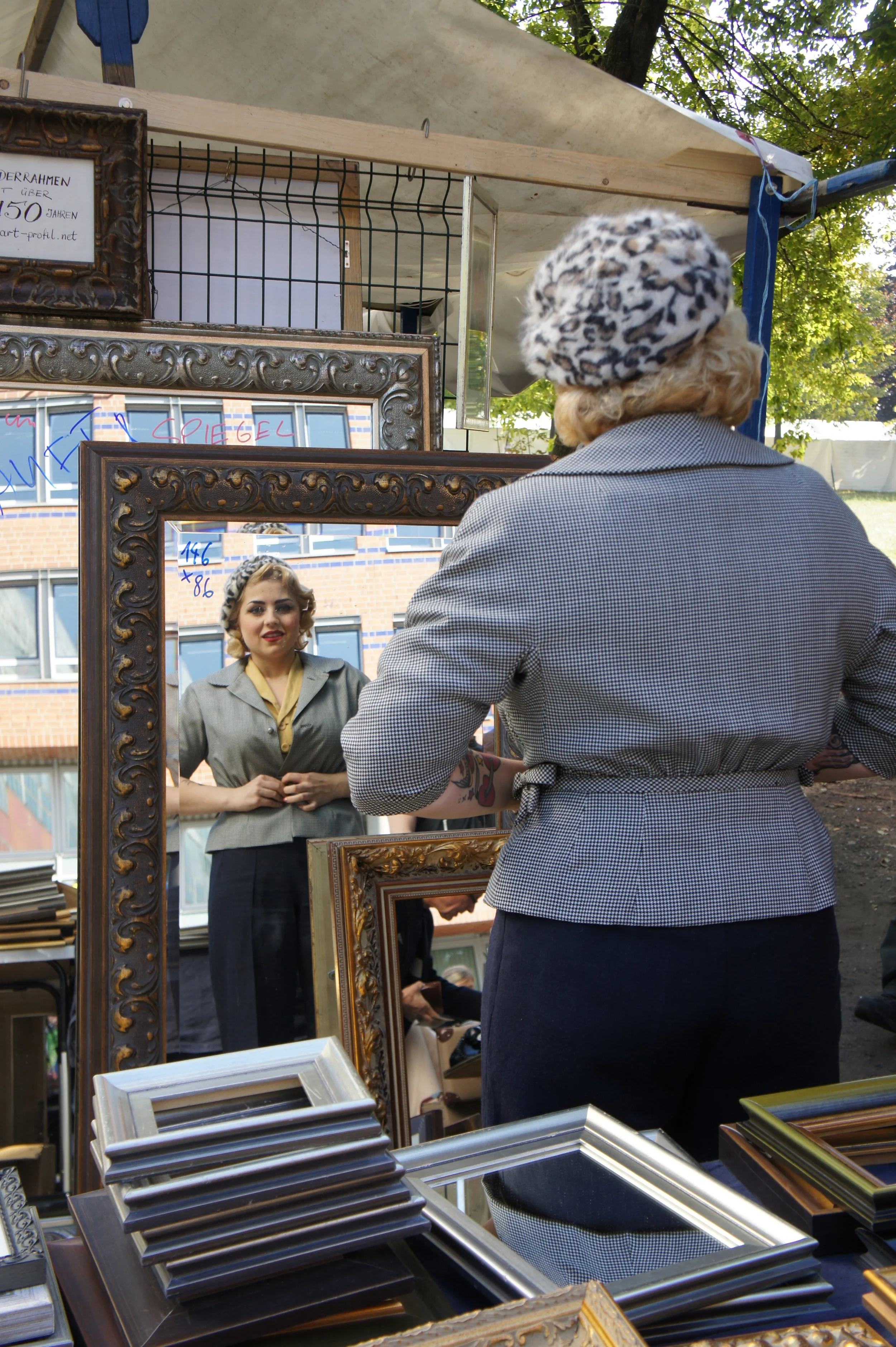 Eine Frau mit blondem Haar schaut in einen Spiegel und trägt einen grauen Blazer. Sie ist auf einem Flohmarkt, die Umkleidekonstruktion ist sichtbar und es gibt mehrere goldene und silberne Rahmen auf einem Tisch im Vordergrund.