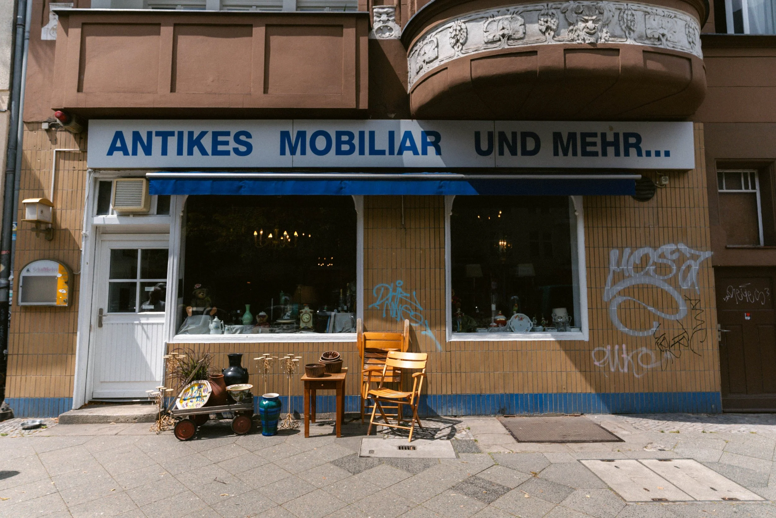 Schaufenster eines Antiquitätengeschäfts mit dem Schild "Antikes Mobiliar und mehr". Davor stehen verschiedene alte Möbelstücke, Vasen und Dekorationsgegenstände.