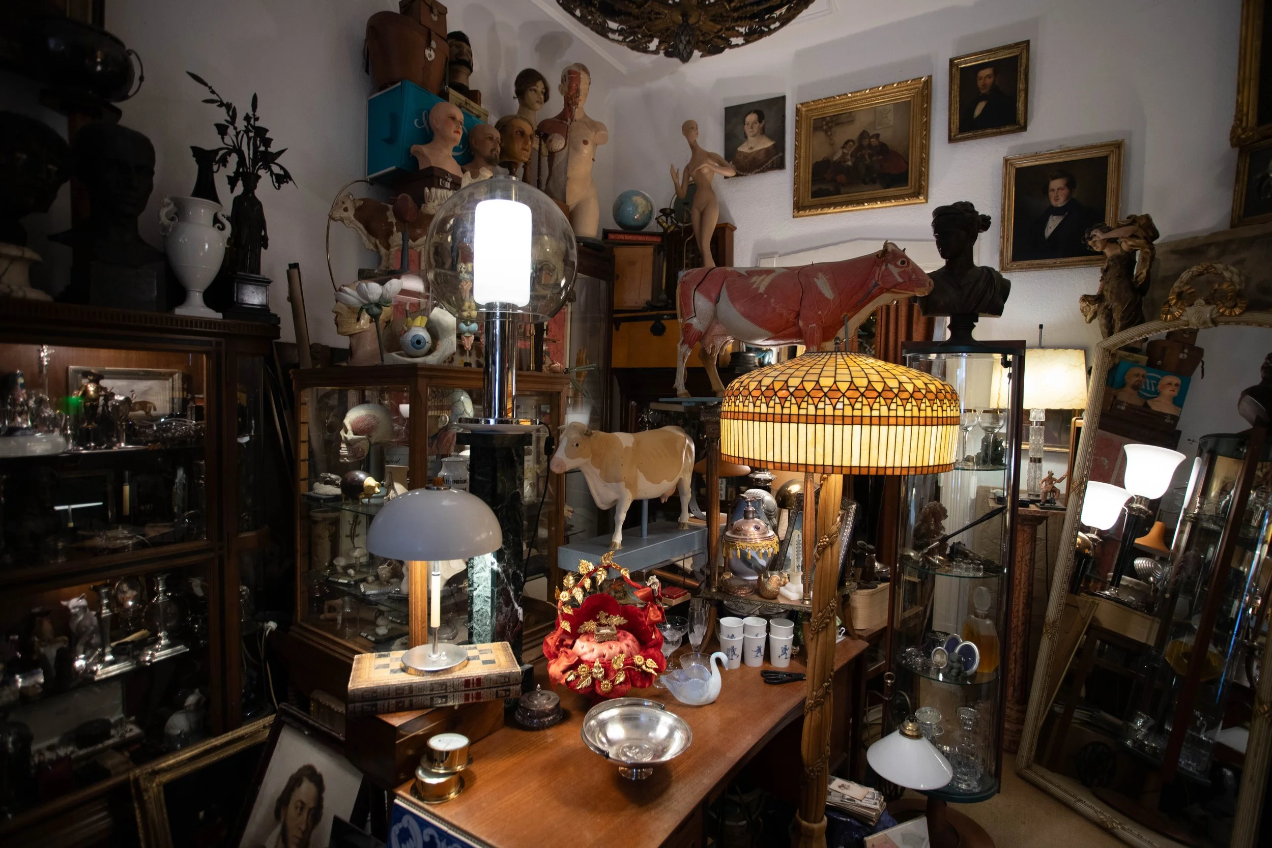 Ein vielschichtiges Arrangement alter Kunstgegenstände, Lampen, Gemälde, Skulpturen, und Dekorationsartikel in einem Raum, vermutlich ein Antiquitätengeschäft oder Lager.