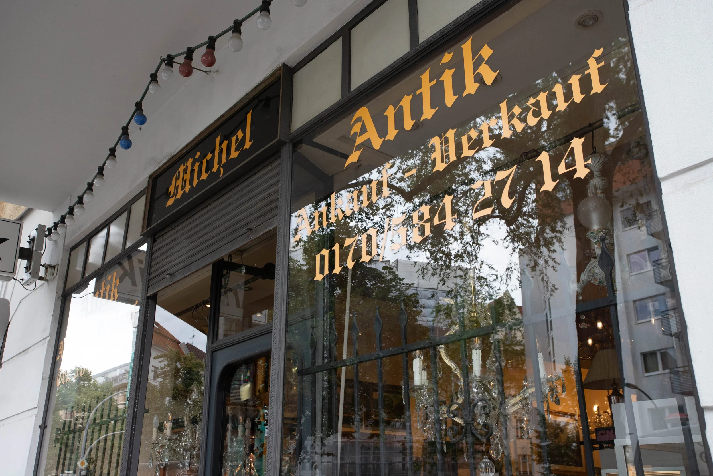 Schaufenster eines Geschäfts mit der Aufschrift 'Kichel Antiquitäten - Antik - Möbel' und einer Telefonnummer, mit einer Gardinenstange und Spiegel im Innenraum, sowie reflektierenden Bäumen und Gebäuden im Fenster