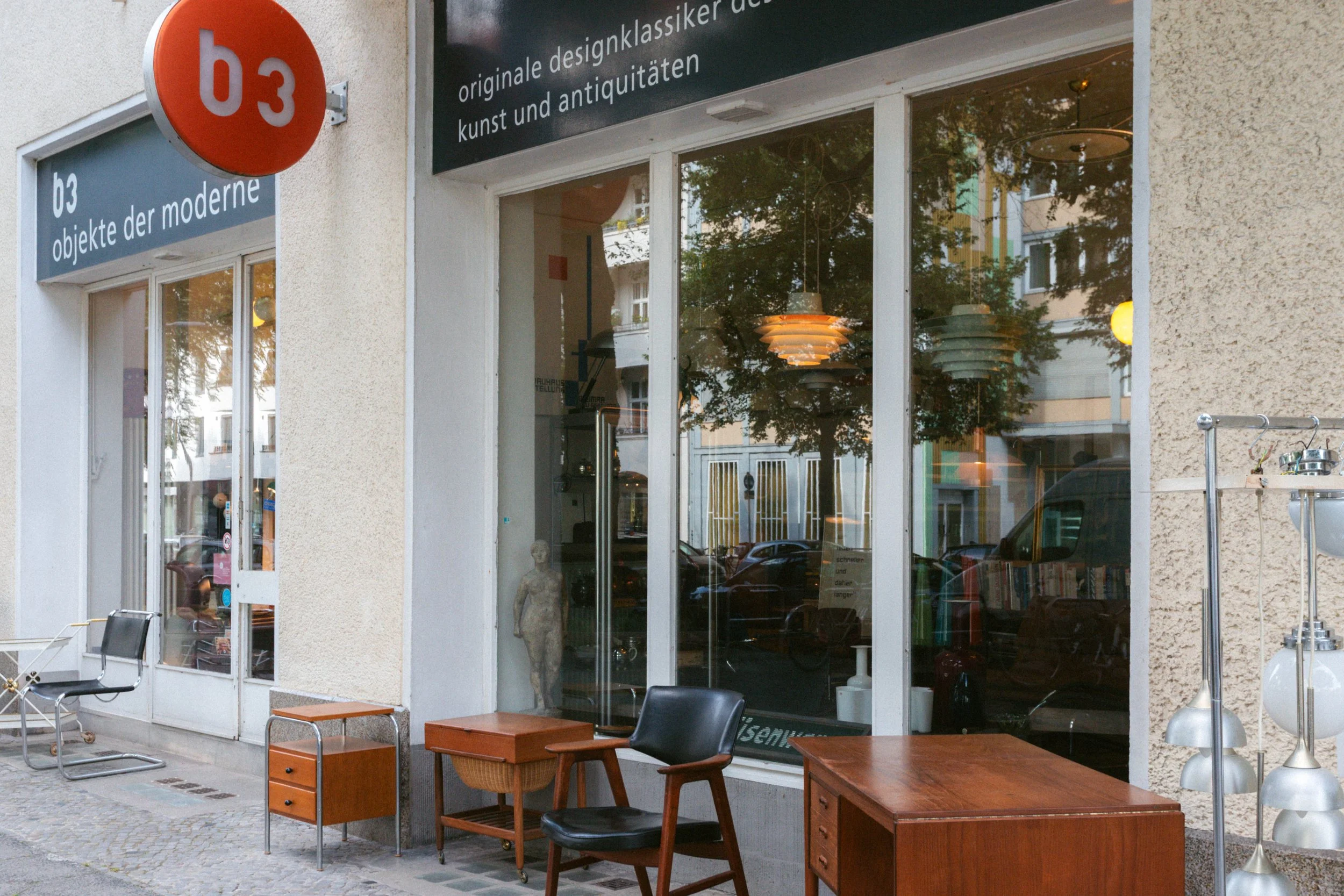 Ein Schaufenster mit antiken Möbeln und Lampen, die im Inneren sichtbar sind, sowie einige moderne Möbelstücke und Dekor auf der Straße davor.