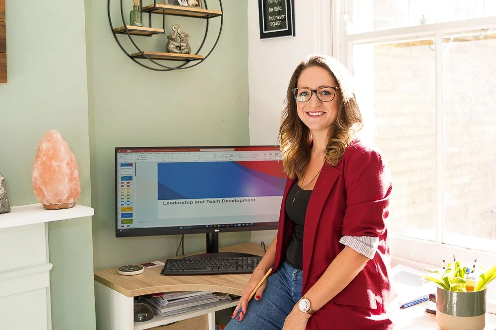Image of Livvy in maroon blazer, leaning on desk, with laptop in the background showing a slide-deck titled "Leadership and Team Development"