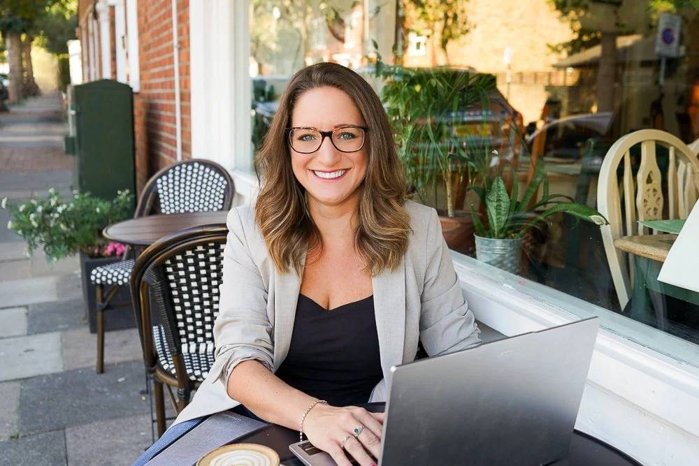 Image of Livvy outside coffee shop with laptop, smiling at camera wearing glasses.