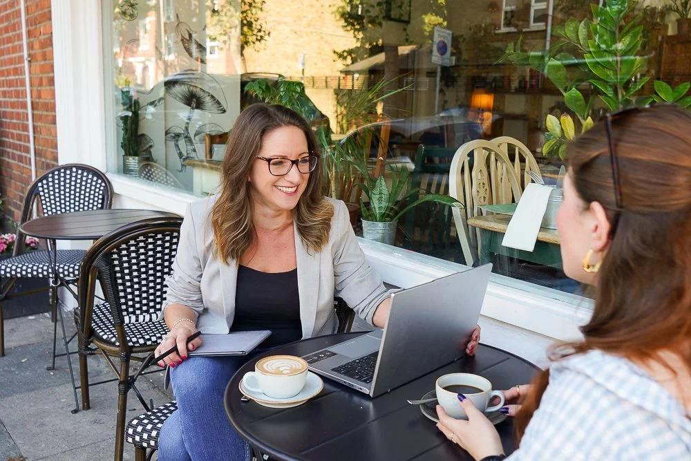 Image of Livvy smiling and talking to a new client. The laptop is positioned on the table between them and there are two coffees in the image.