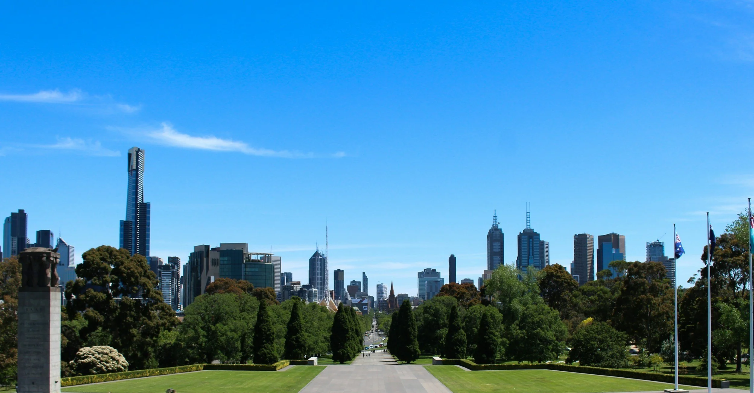 View of a city skyline with tall skyscrapers and a large park with trees and flags in the foreground, under a clear blue sky.