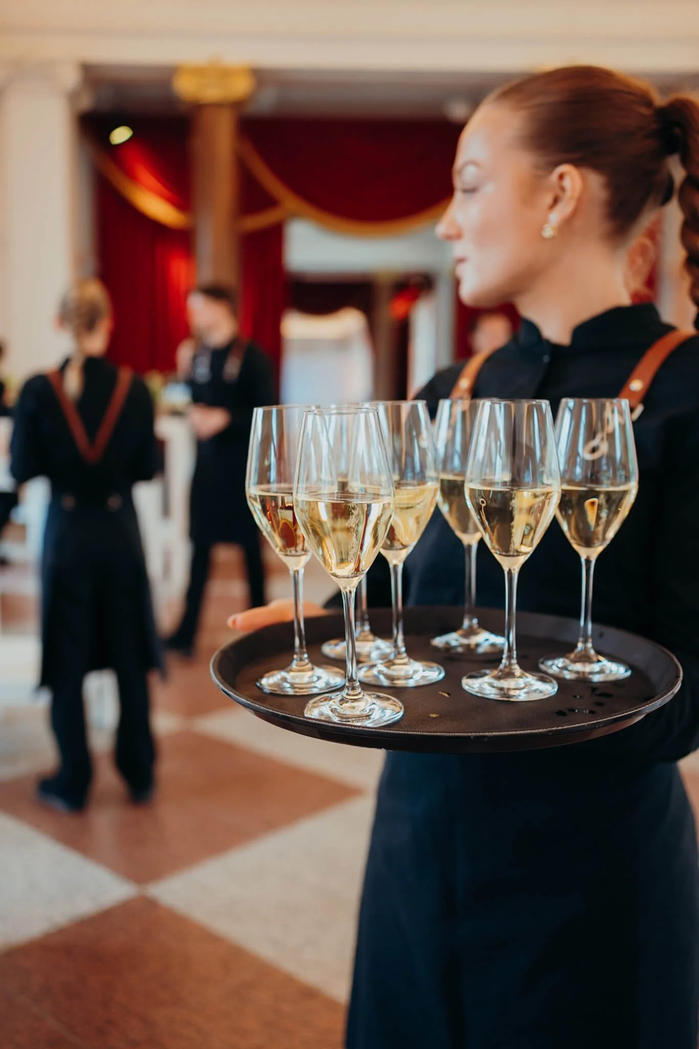 A waitress holding a tray of six glasses of white wine in a restaurant or event space.