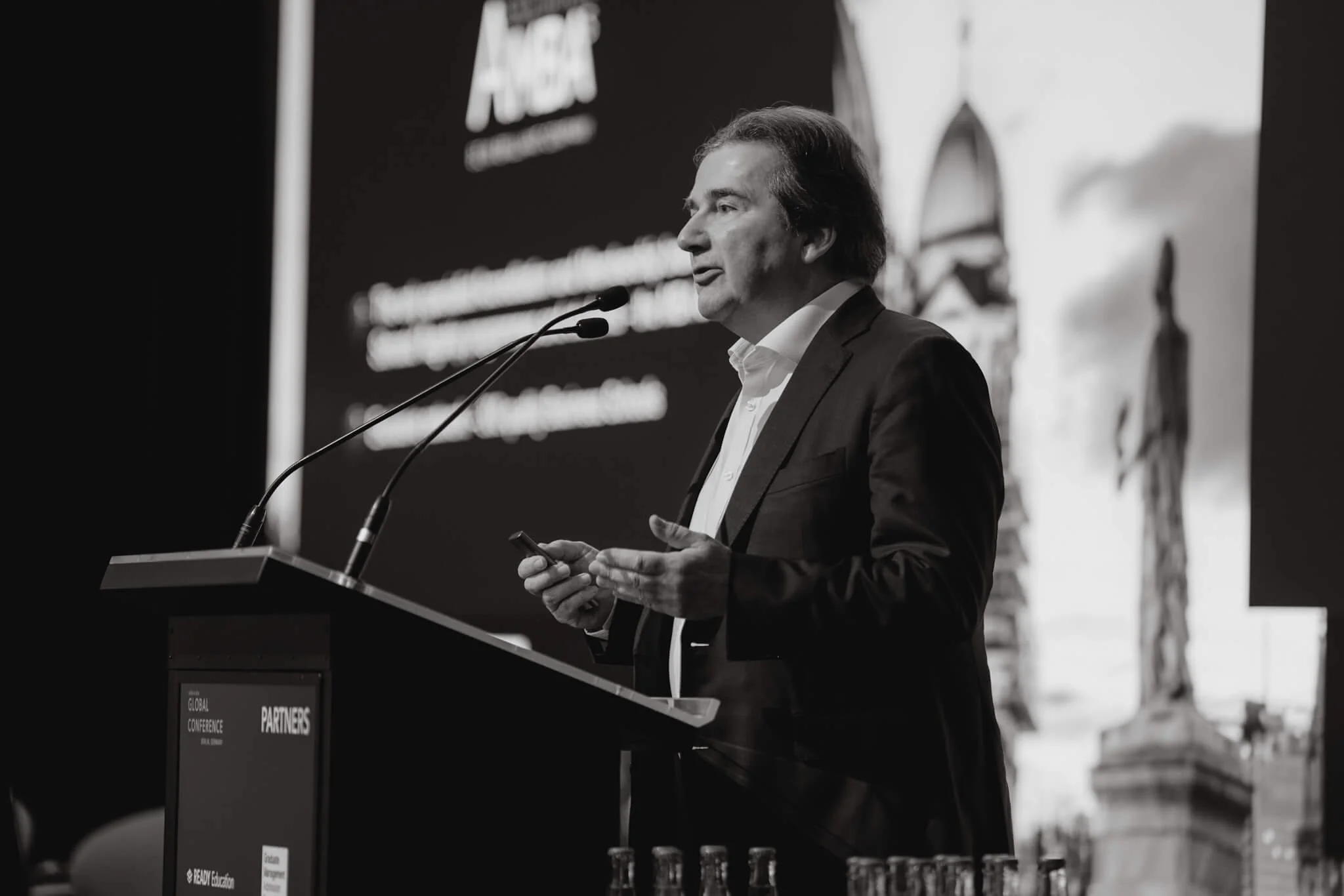 A man in a suit speaking at a podium during a conference, holding a microphone in one hand and a remote control in the other, with a large screen displaying a presentation behind him.