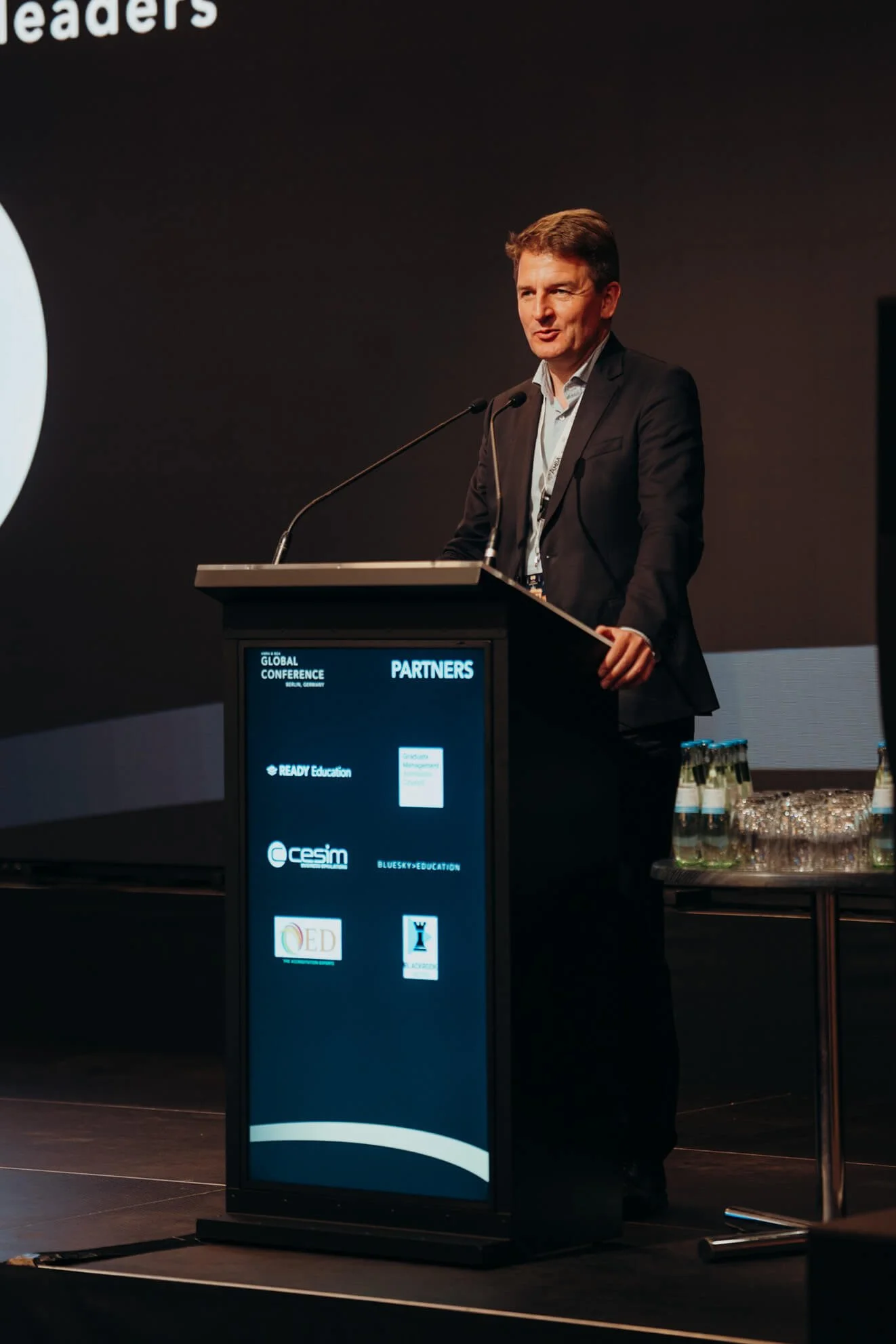 A man in a suit standing at a podium speaking during a conference with a dark background and a screen displaying the conference's partner logos.