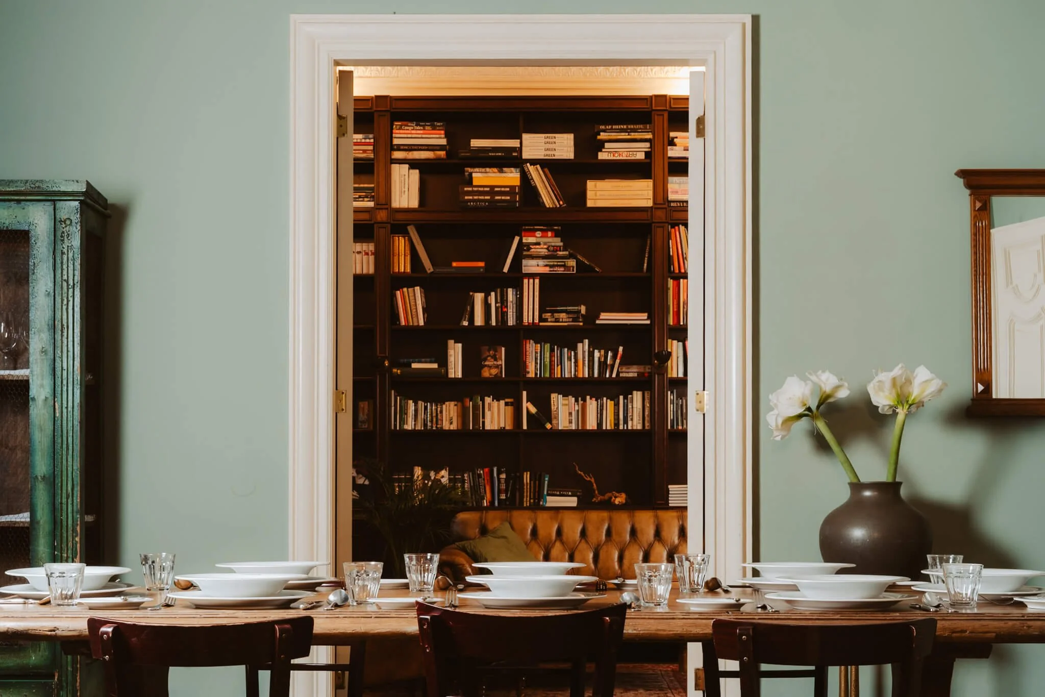 A dining room view through a window opening with a view of a bookshelf filled with books in the background. The foreground shows a wooden dining table set with white bowls, plates, and clear glasses. To the right, there is a dark vase with white flow